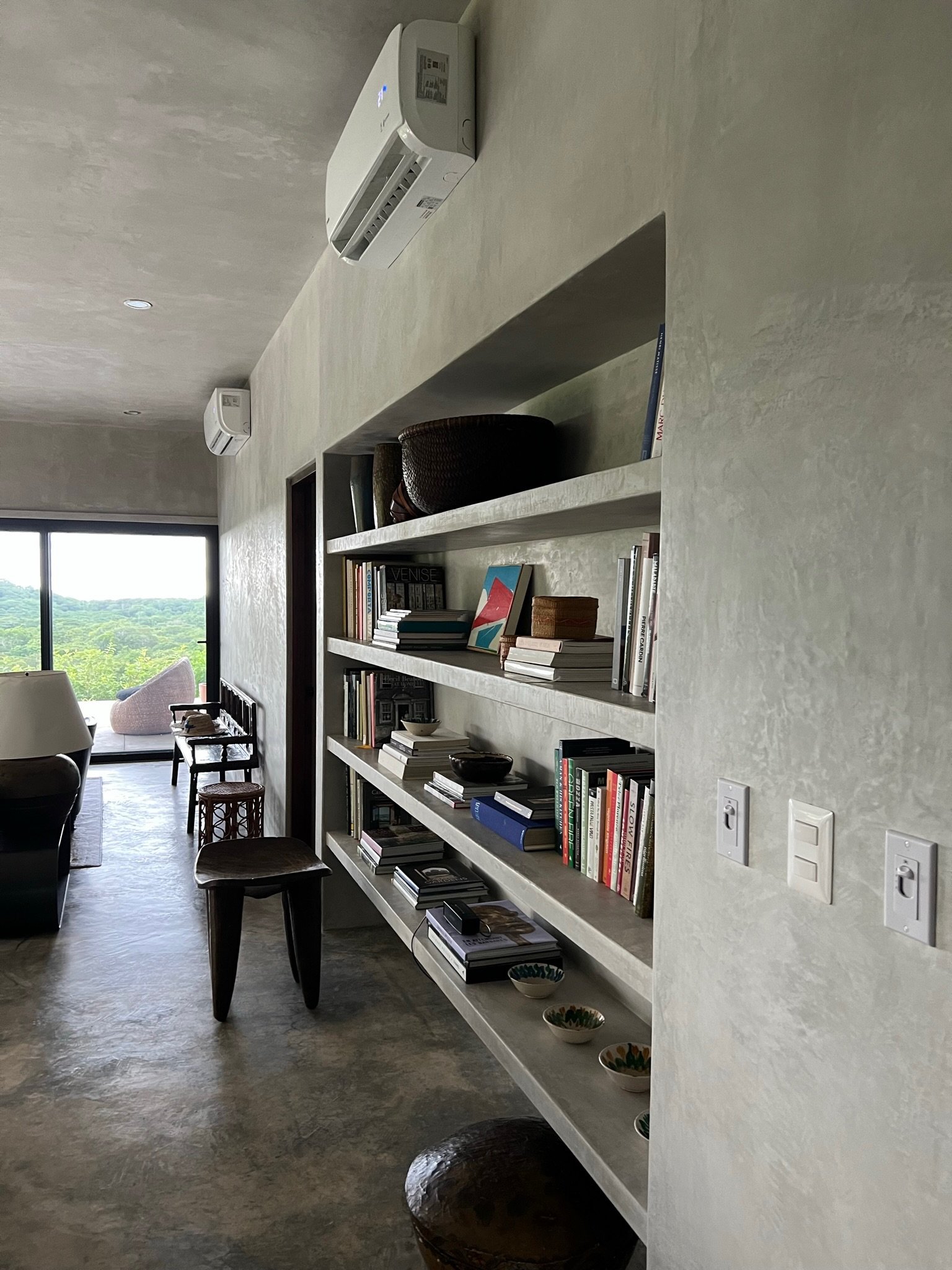 Living room with built-in bookshelves filled with books and decorative objects, a dark wooden stool, a window with a view of greenery outside, and air conditioning units mounted on the wall.