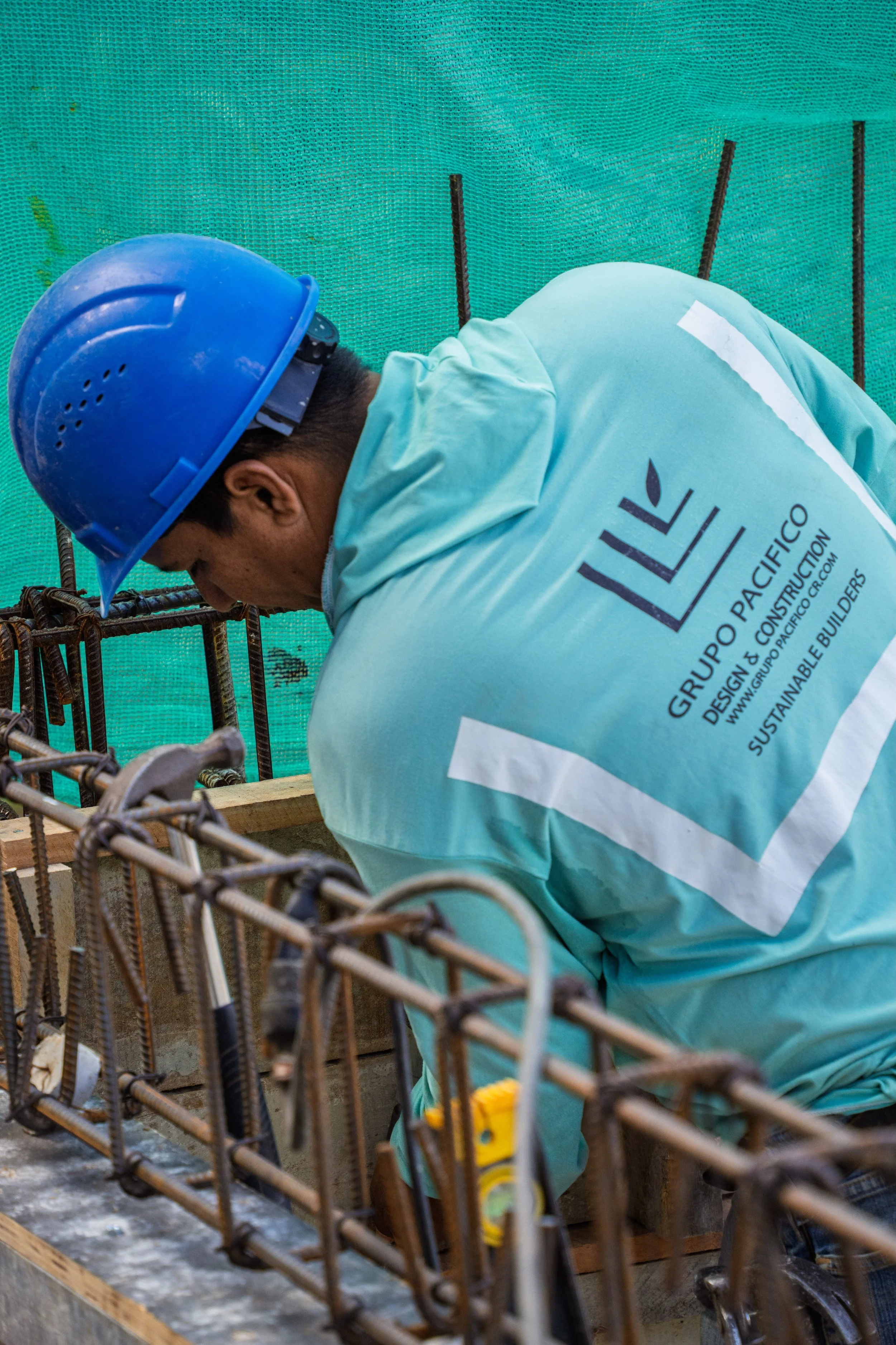 A construction worker wearing a blue safety helmet and a teal jacket labeled Grupo Pacifico Design & Construction is working on a concrete structure with steel rebar.