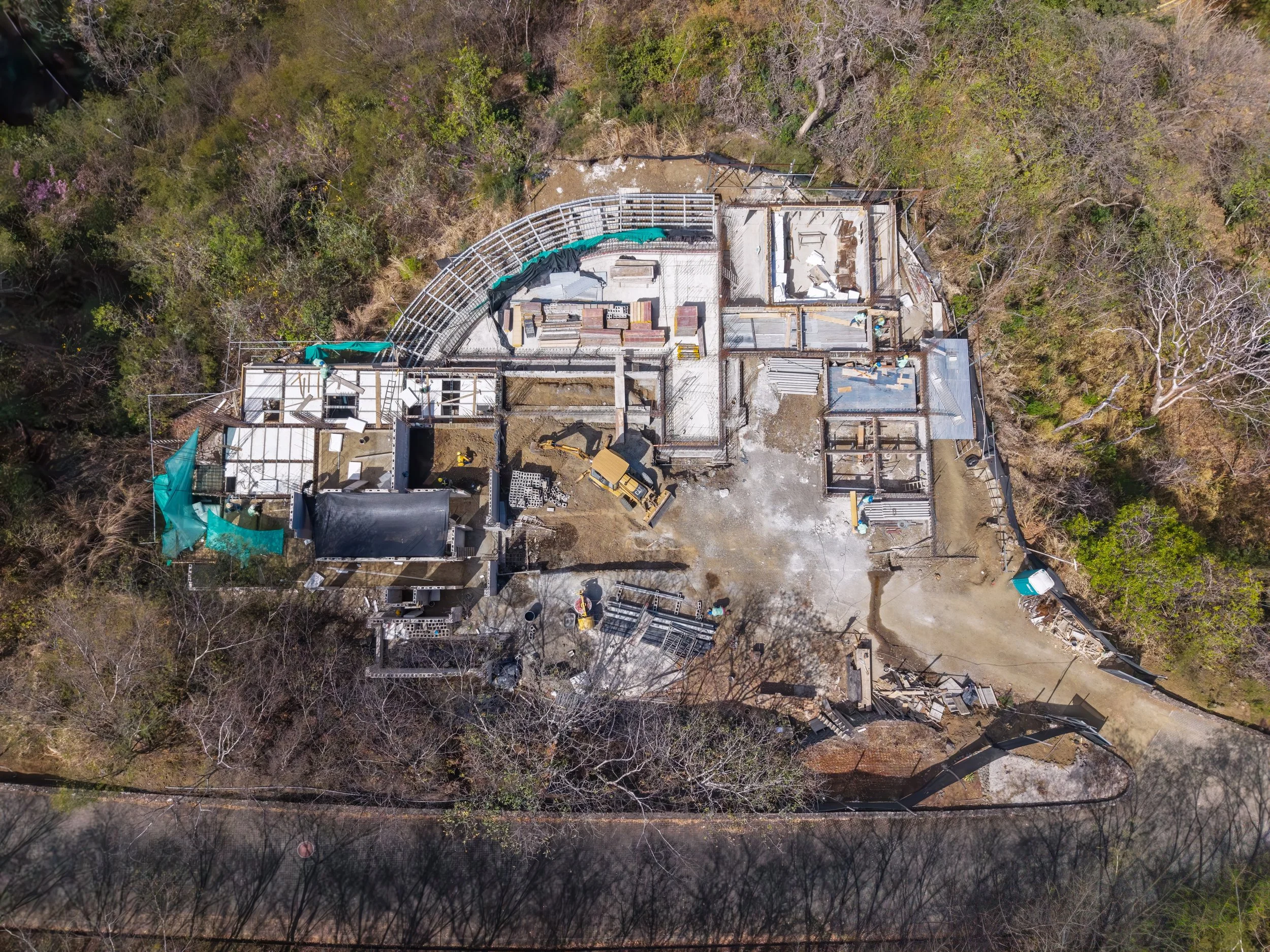 An aerial view of a house under construction with visible framework, construction equipment, and materials surrounded by trees.