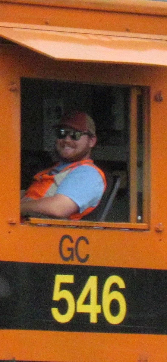 A man wearing sunglasses, a red cap, and an orange safety vest sitting at a control panel of a train or tram with the markings 'GC 546'. The background shows a dark interior.