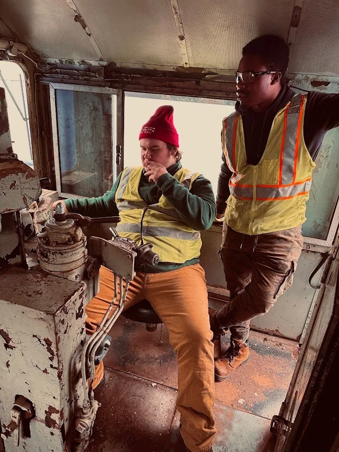 Two workers in a control room of a train or tram, one seated and operating controls, the other standing nearby, both wearing high-visibility vests.