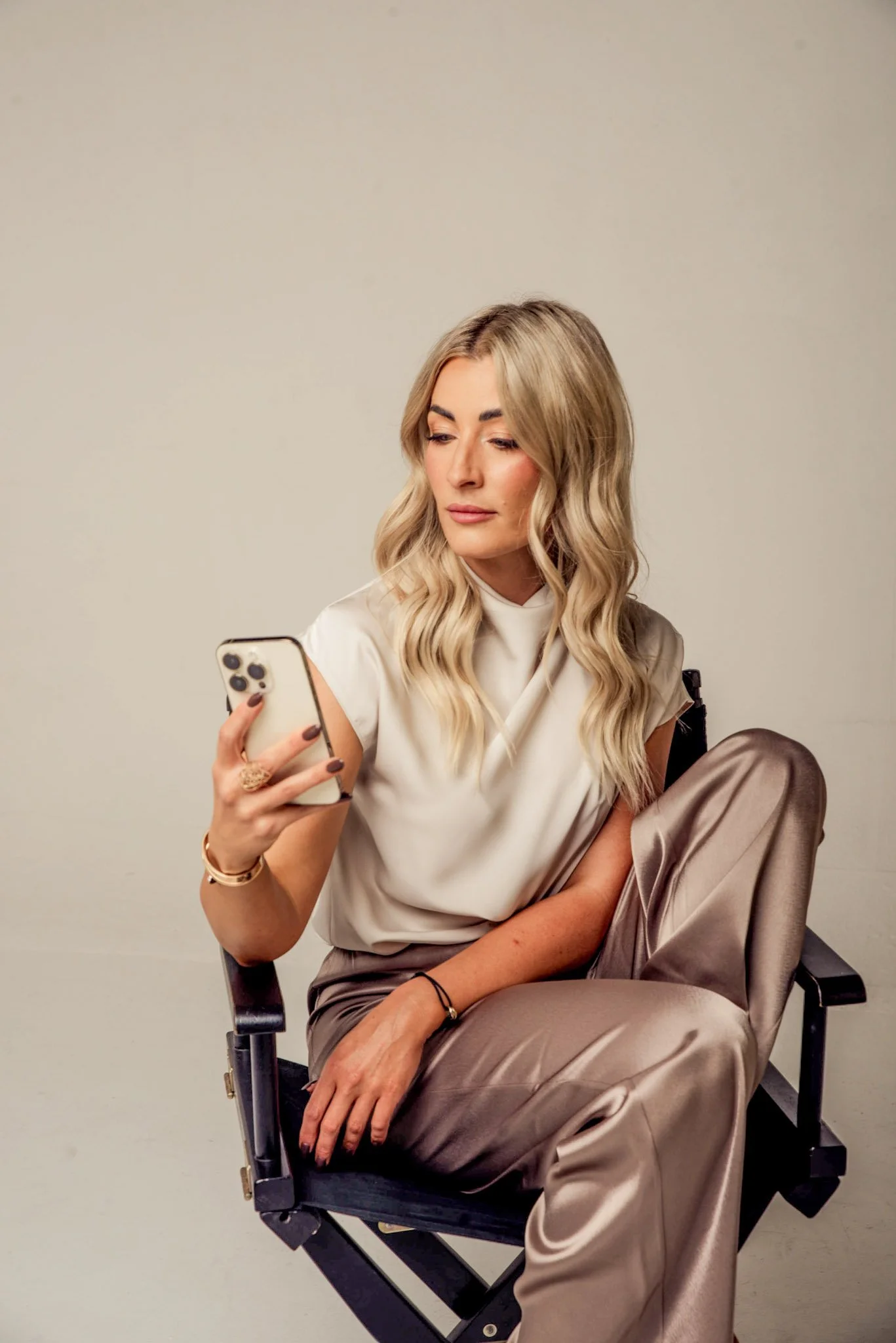A woman with long wavy blonde hair wearing a satin beige top and pants, sitting in a director's chair, looking at her smartphone against a plain, light-colored background.