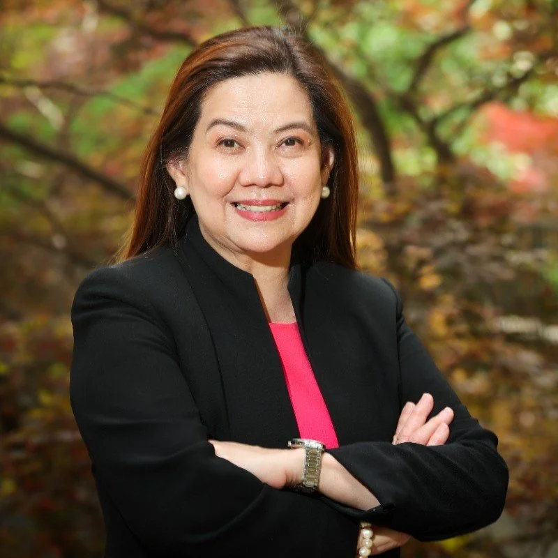 A professional woman with long dark hair smiling, standing outdoors with blurred autumn leaves in the background, wearing a black blazer, pink top, pearl earrings, and a watch.