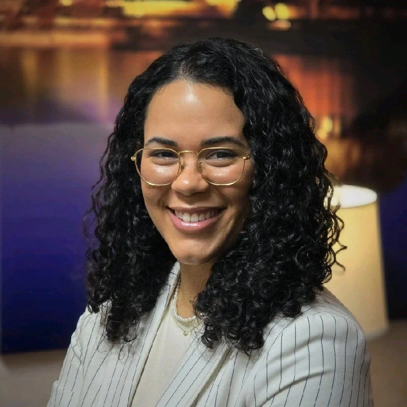A woman with curly black hair wearing glasses and a pinstripe blazer, smiling in an indoor setting.