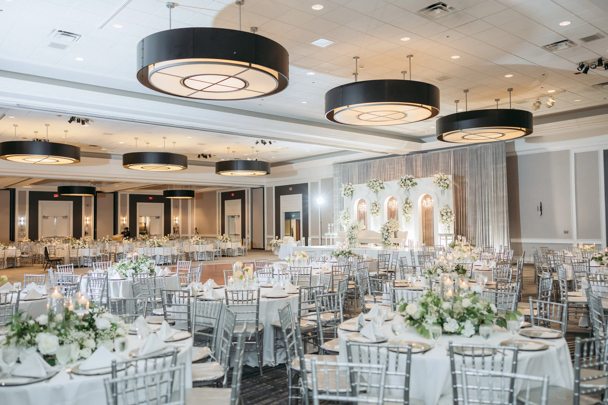 Elegant banquet hall decorated for a wedding reception with numerous round tables covered in white tablecloths, floral centerpieces, silver chairs, and a floral backdrop on stage with curtains.