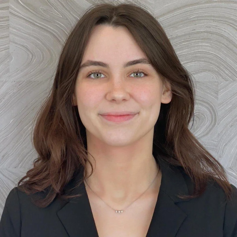 A young woman with shoulder-length brown hair, green eyes, and light skin, wearing a black blazer and a delicate necklace, smiling against a textured gray background.