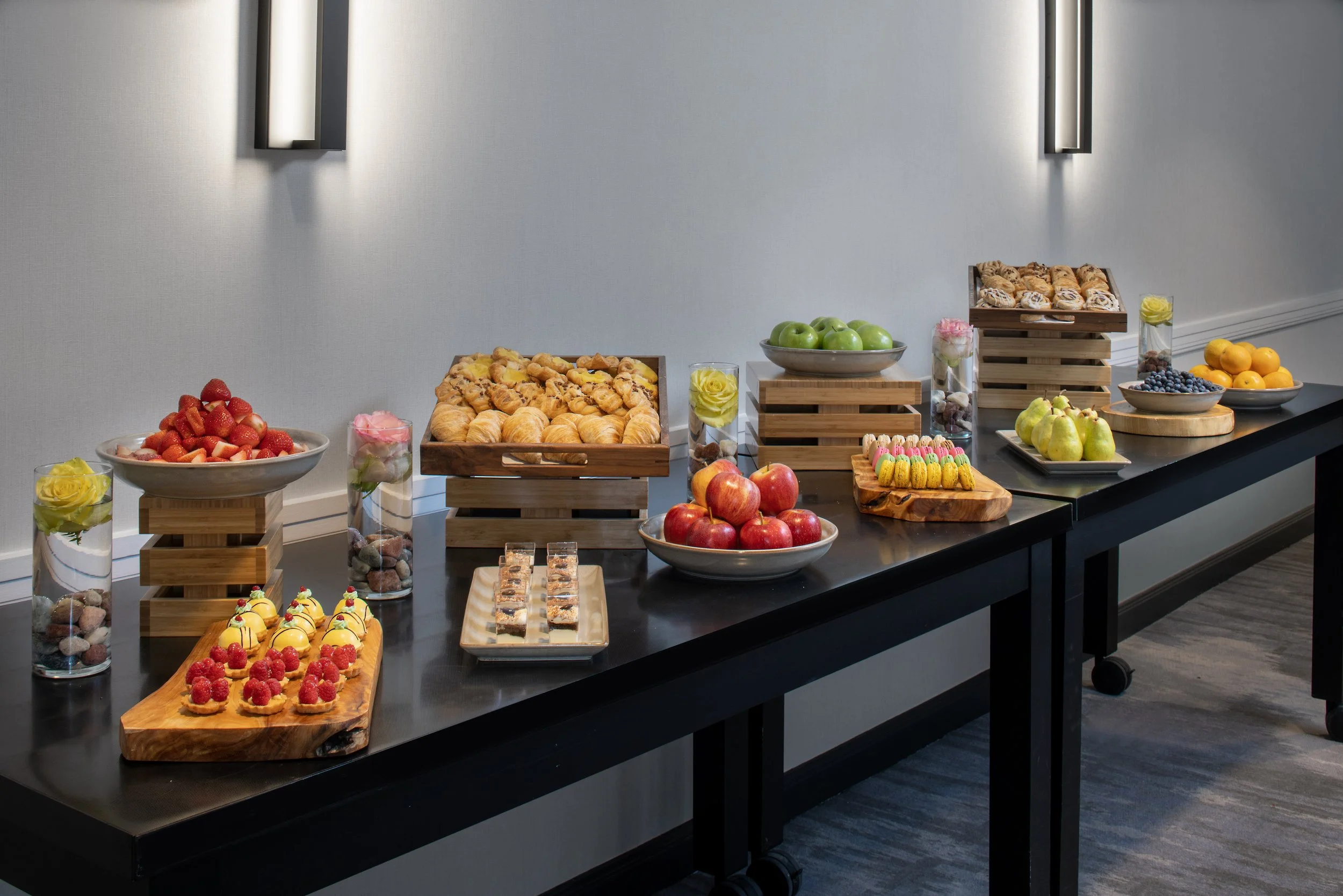 Assorted fresh fruit and pastries arranged on a black table with decorative centerpieces and lighting.