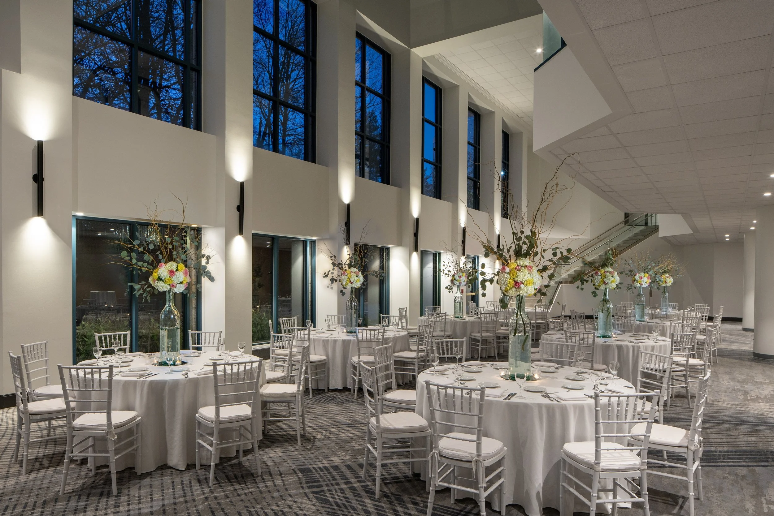 An elegant event space with round tables covered in white tablecloths, each decorated with tall glass vases filled with floral arrangements, overlooking a large windowed wall with a night sky in the background.