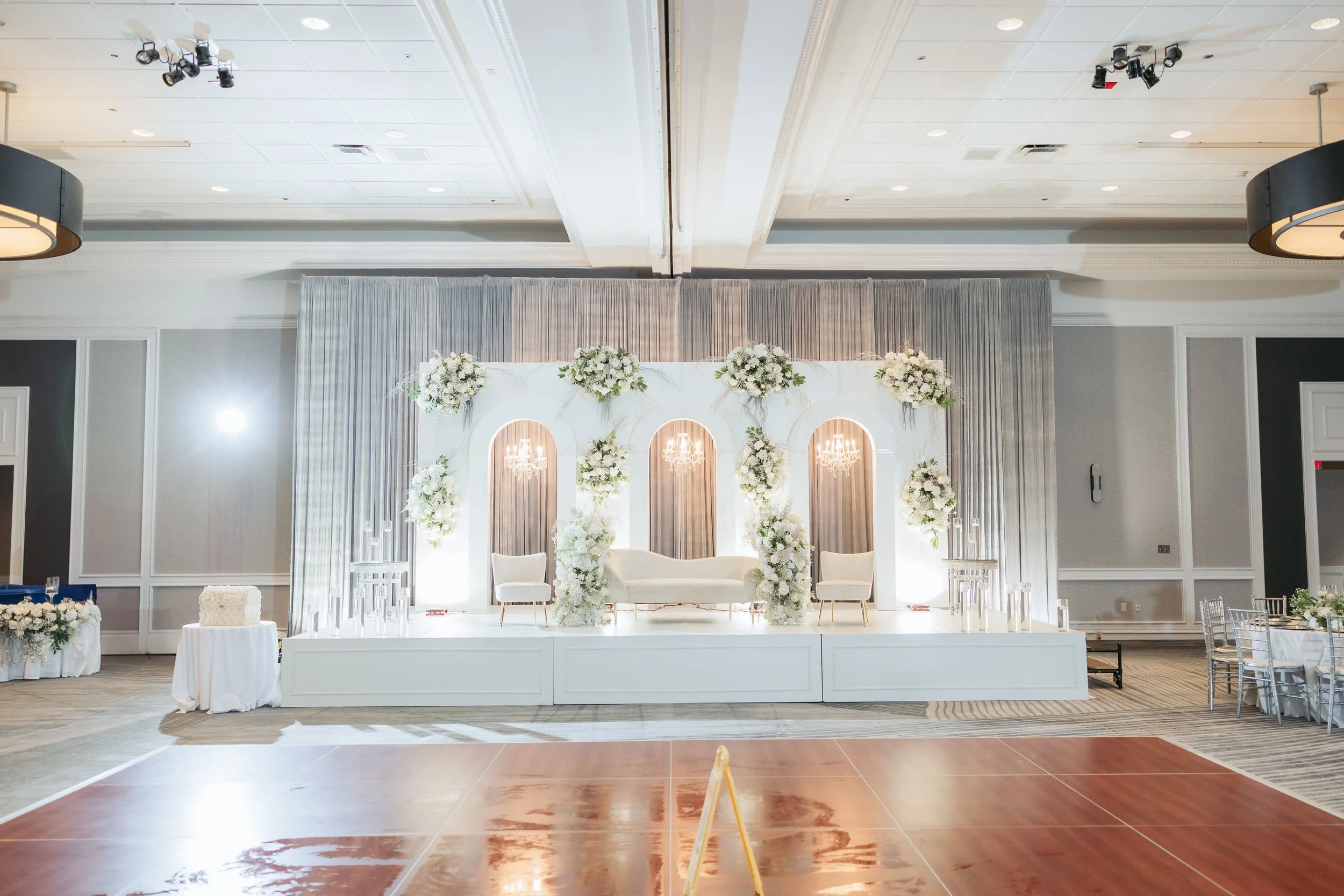 A decorated wedding reception stage with white flowers, chandeliers, and chairs in elegant setting.