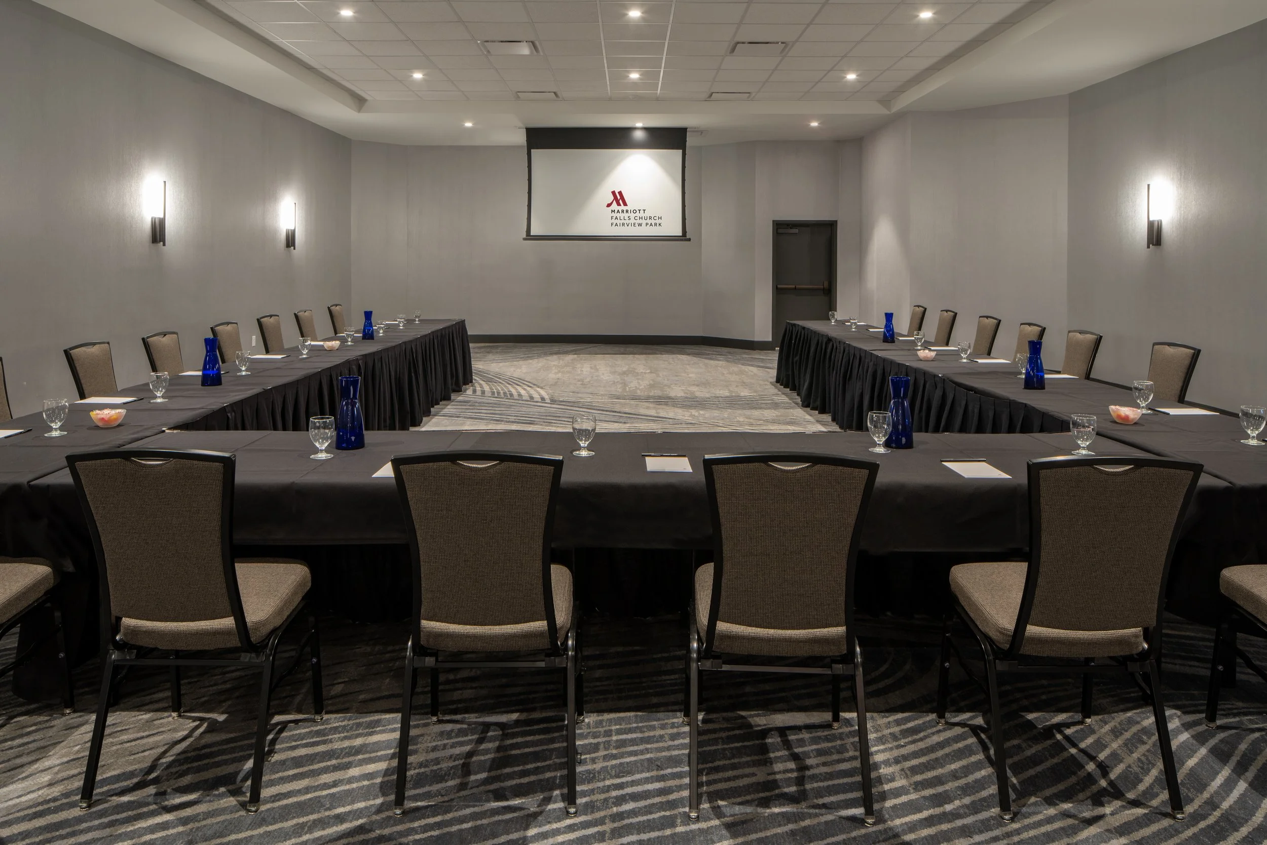 Empty conference room with U-shaped table setup, black tablecloths, brown chairs, blue water pitchers, glasses, and small bowls, in a hotel ballroom with gray walls and a projector screen displaying the Marriott logo.