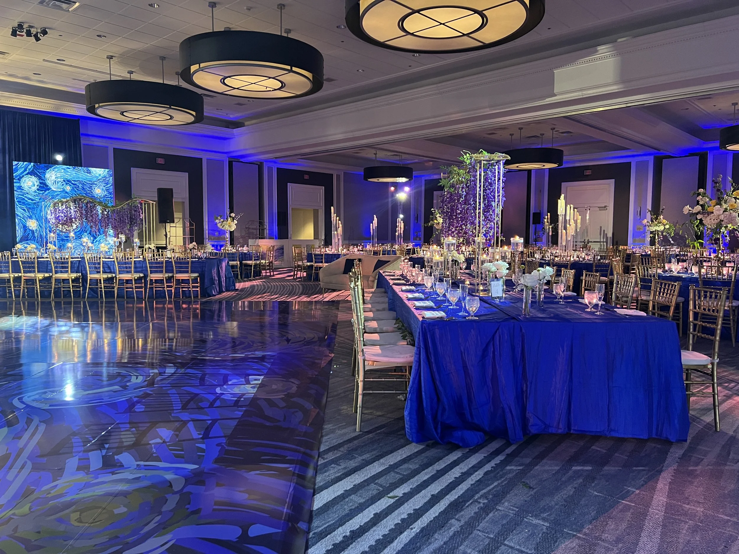 Elegantly decorated banquet hall with round tables and long rectangular tables draped in deep blue cloths, adorned with floral centerpieces and candles, illuminated by blue accent lighting, with a large projection screen displaying a starry sky paint