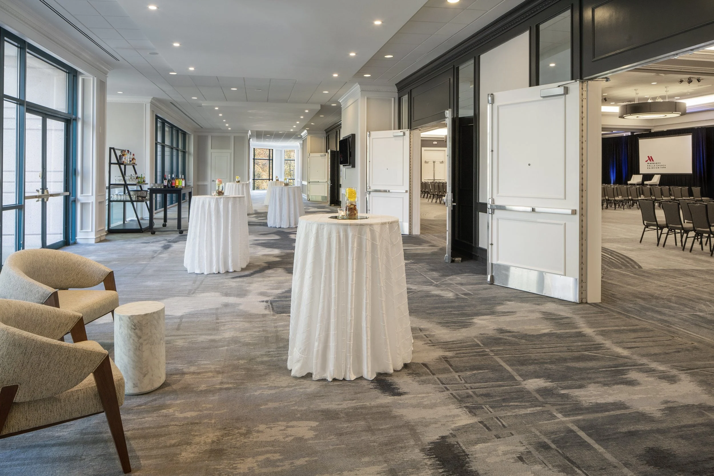 Empty event space with high tables covered in white cloths, a seating area with beige chairs and a small round table, and a bar area with bottles near large glass windows. Chairs are arranged in rows in another room visible through the open doors.