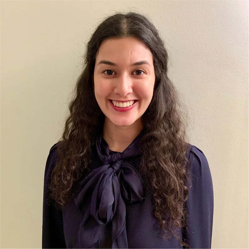 A young woman with long, curly dark hair smiling at the camera, wearing a dark blouse with a bow tie at the collar, standing against a plain light-colored wall.