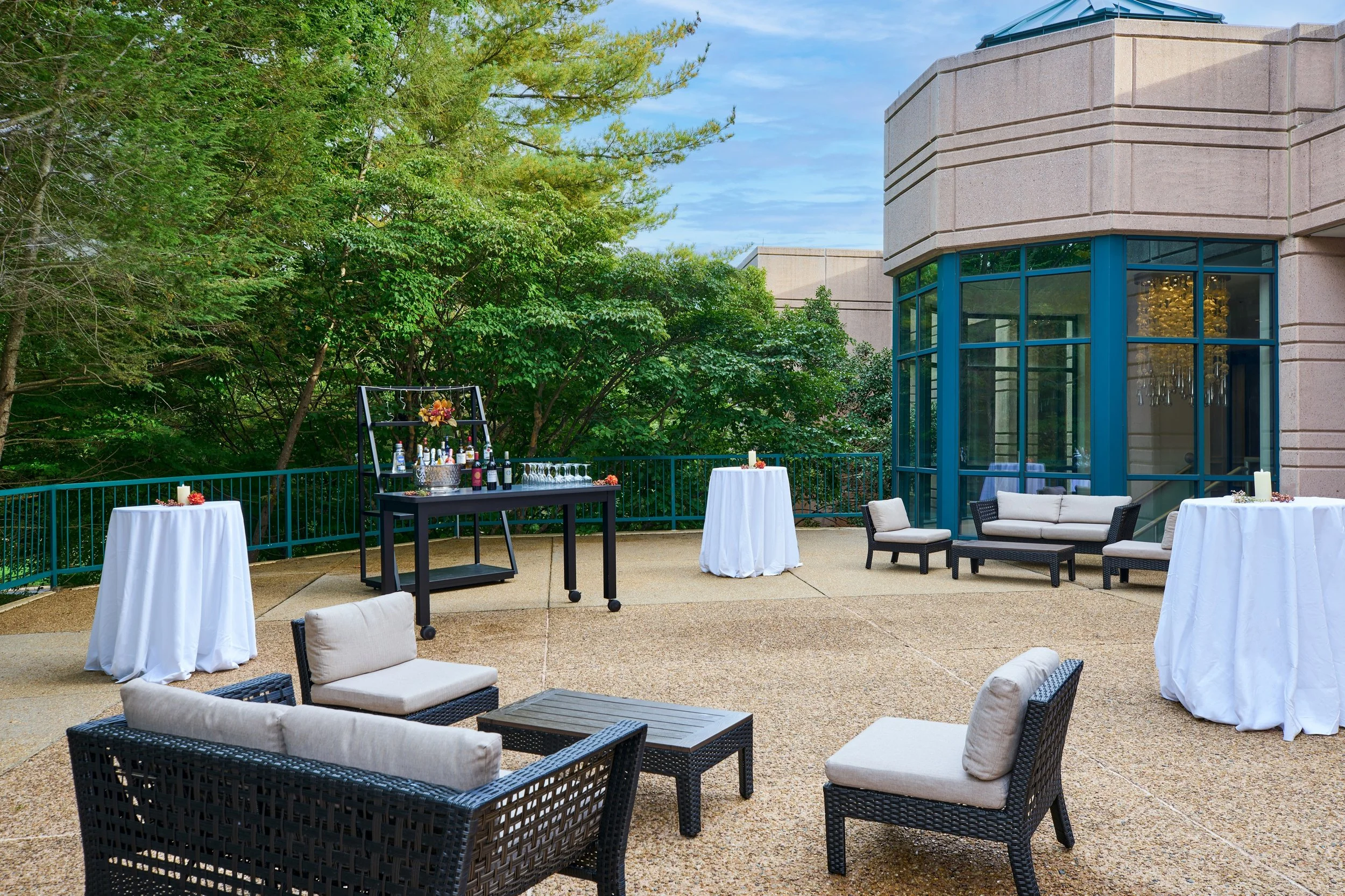 An outdoor patio setup featuring high-top tables with white tablecloths, a black bar cart with bottles and glasses, a beige seating area with cushioned chairs and a sofa, surrounded by lush green trees, with a modern building with large blue-framed w