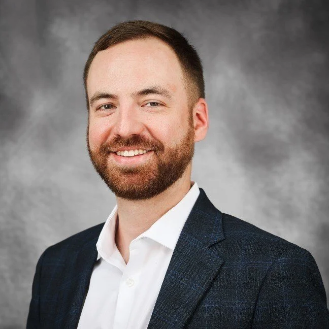 A man with short brown hair and a beard smiling, wearing a navy blazer and white dress shirt against a gray background.