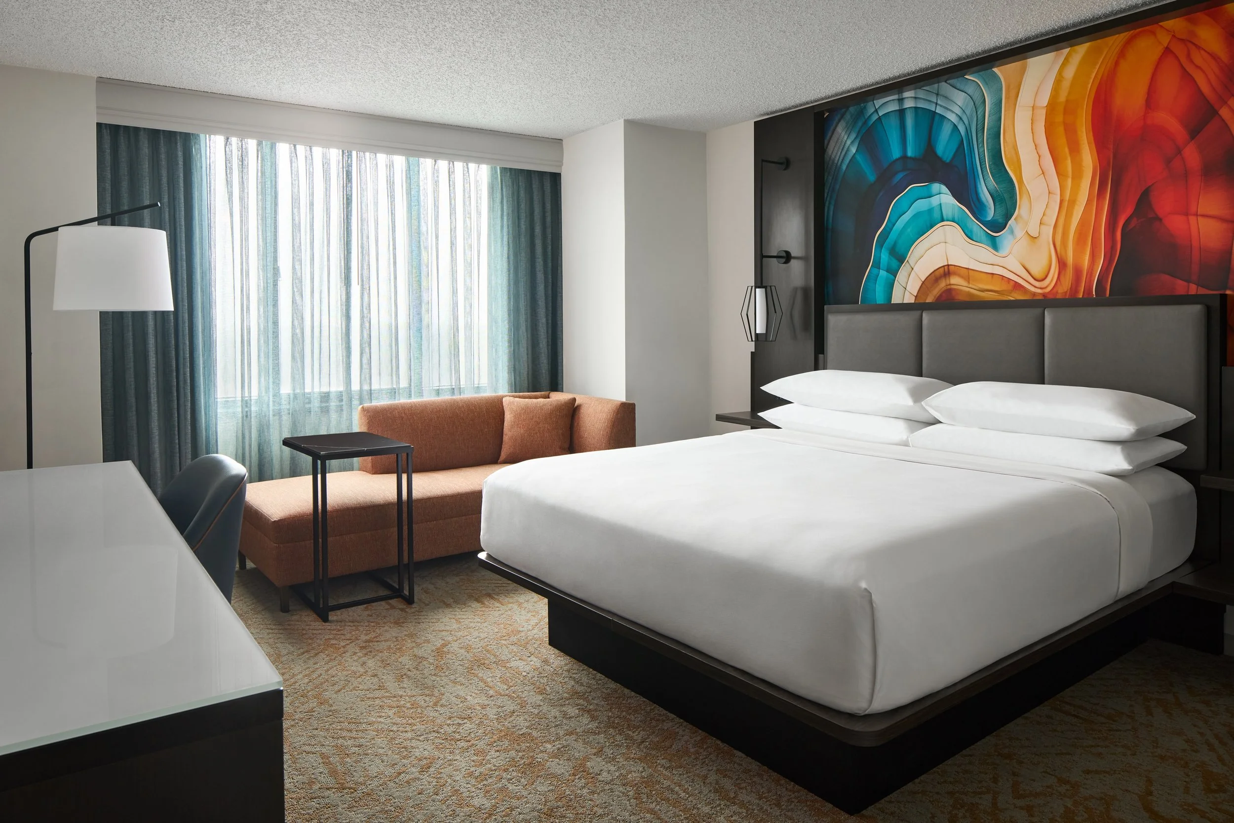 A hotel room with two white beds, a nightstand, table lamps, a chair, a desk with a black chair, and a large window with a view of trees and sky.