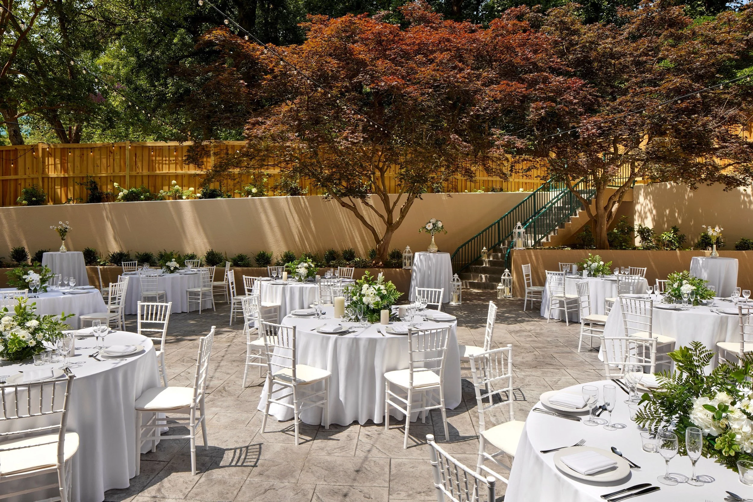 Outdoor wedding or event setup with round tables covered in white tablecloths, white chairs, and floral centerpieces, surrounded by trees and a wooden fence.