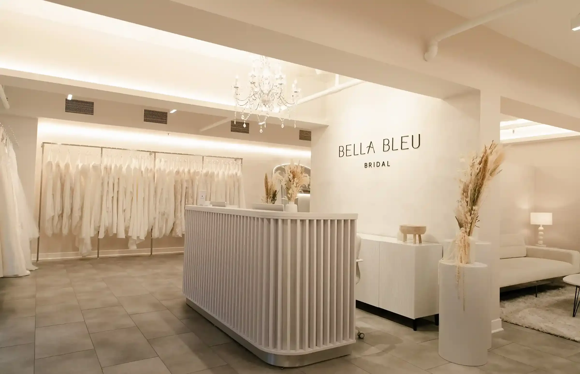Interior of Bella Bleu bridal store with white wedding dresses on racks, a chandelier, and minimalist decor.