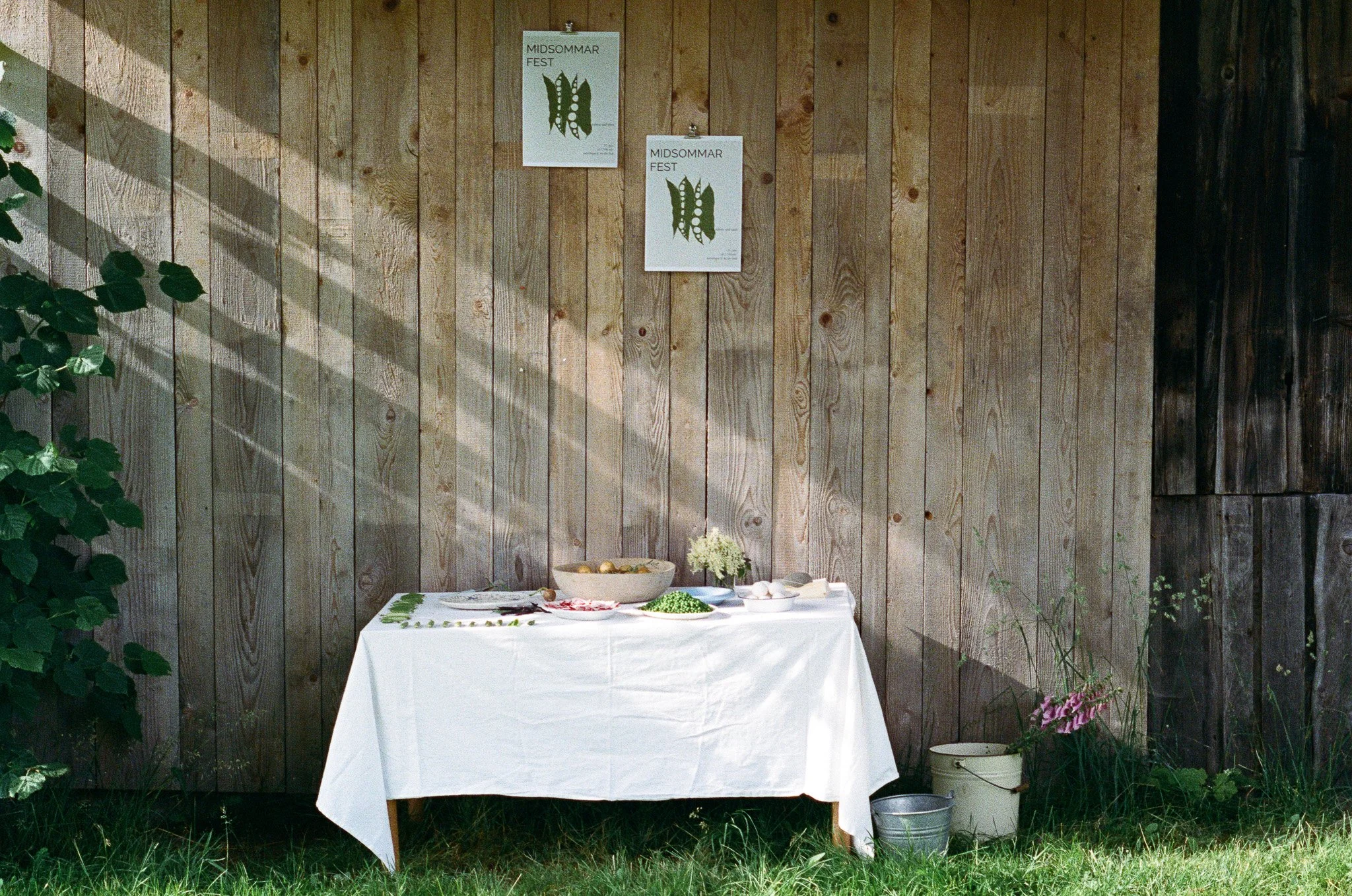A table covered with a white tablecloth with various bowls of food and a small flower arrangement, set against a wooden fence with posters that read 'MIDSOMMAR FEST' in an outdoor setting.