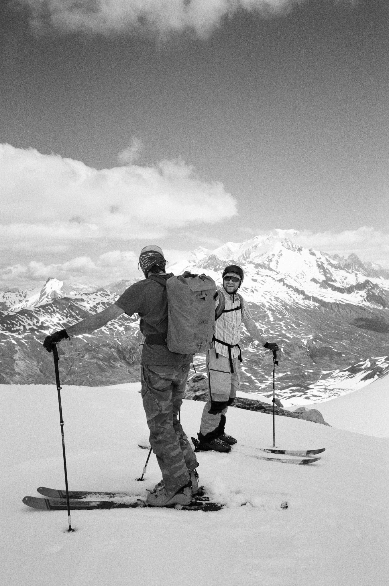 Two people snow skiing on a mountain with snow-covered peaks and clouds in the sky.