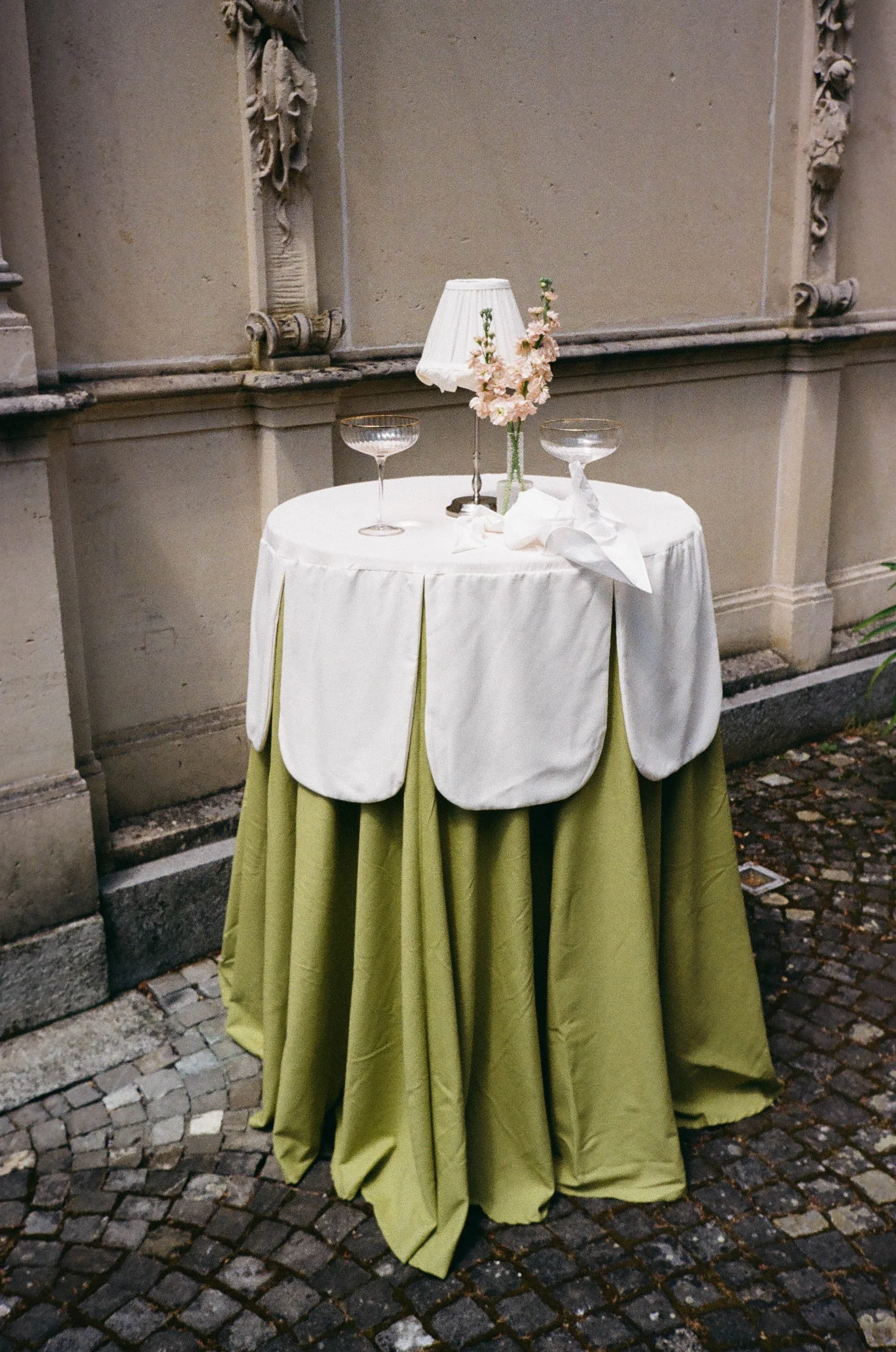 A wedding table with white and green tablecloth, positioned outside on cobblestone pavement against a beige stone wall captured by Hochzeitsfotograf in Zürich Schweiz