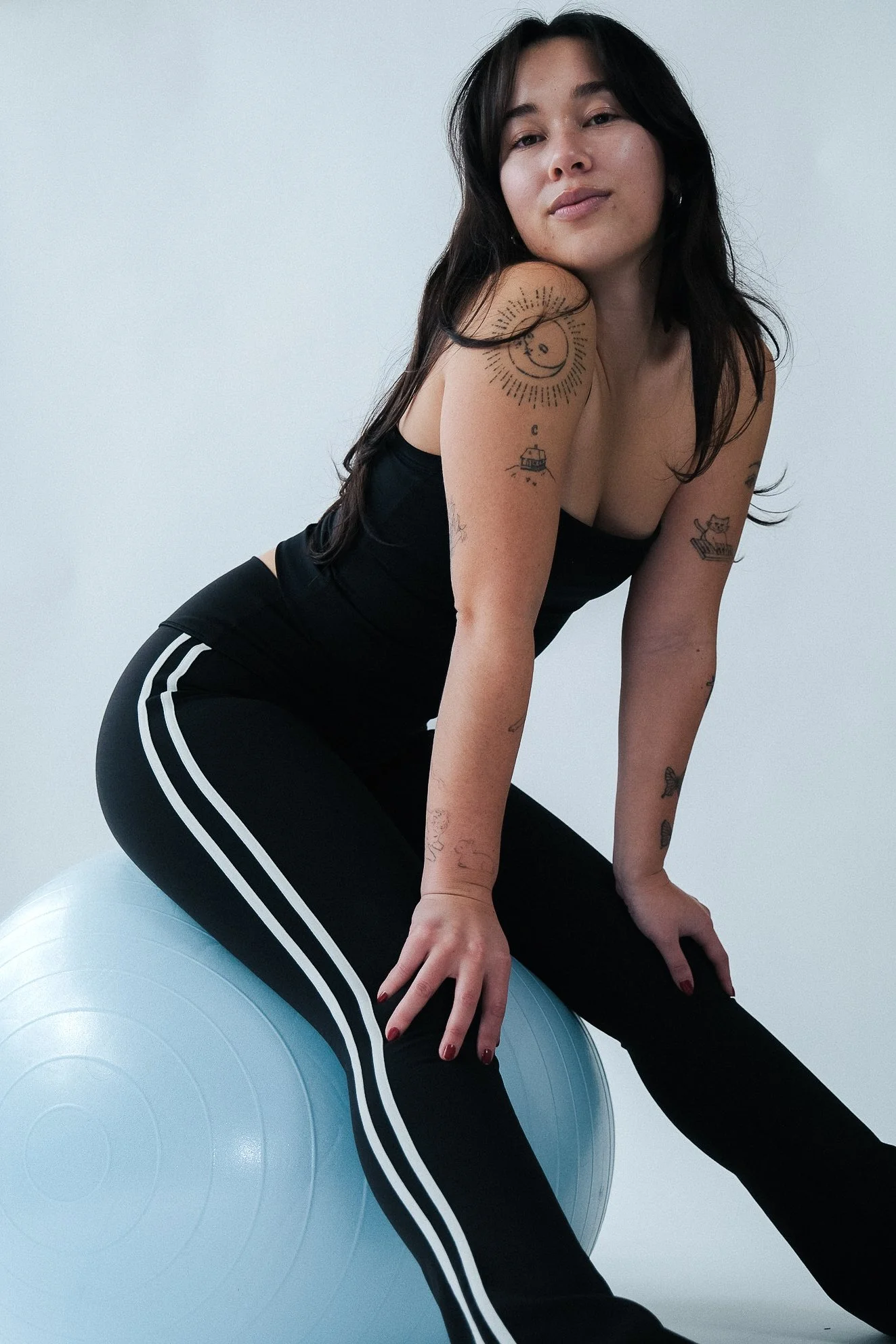 Woman in black athletic wear with white stripes pose sitting on a blue exercise ball against a plain white wall