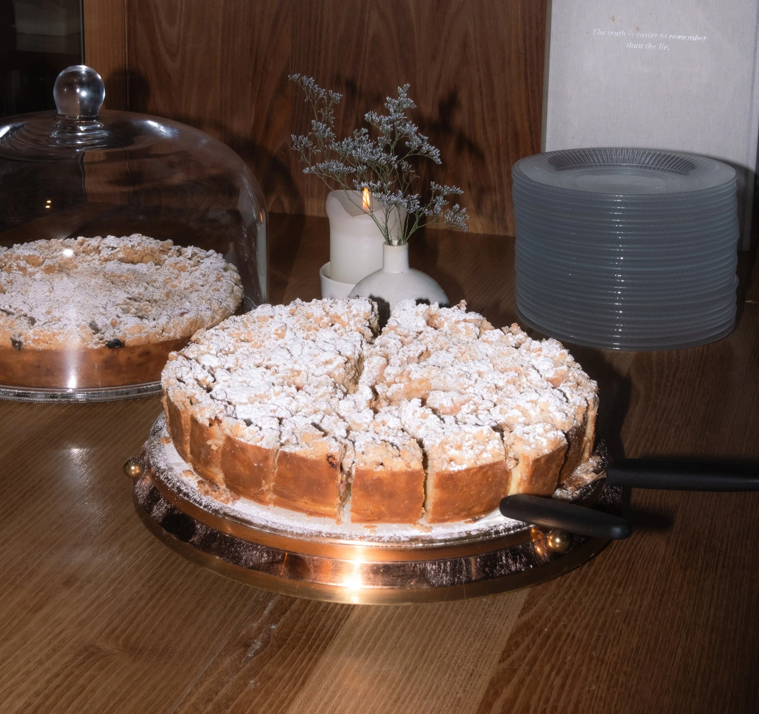 A sliced cake dusted with powdered sugar on a cake stand, with servings cut and a cake cover nearby, on a wooden table with a small vase containing flowers and a candle.