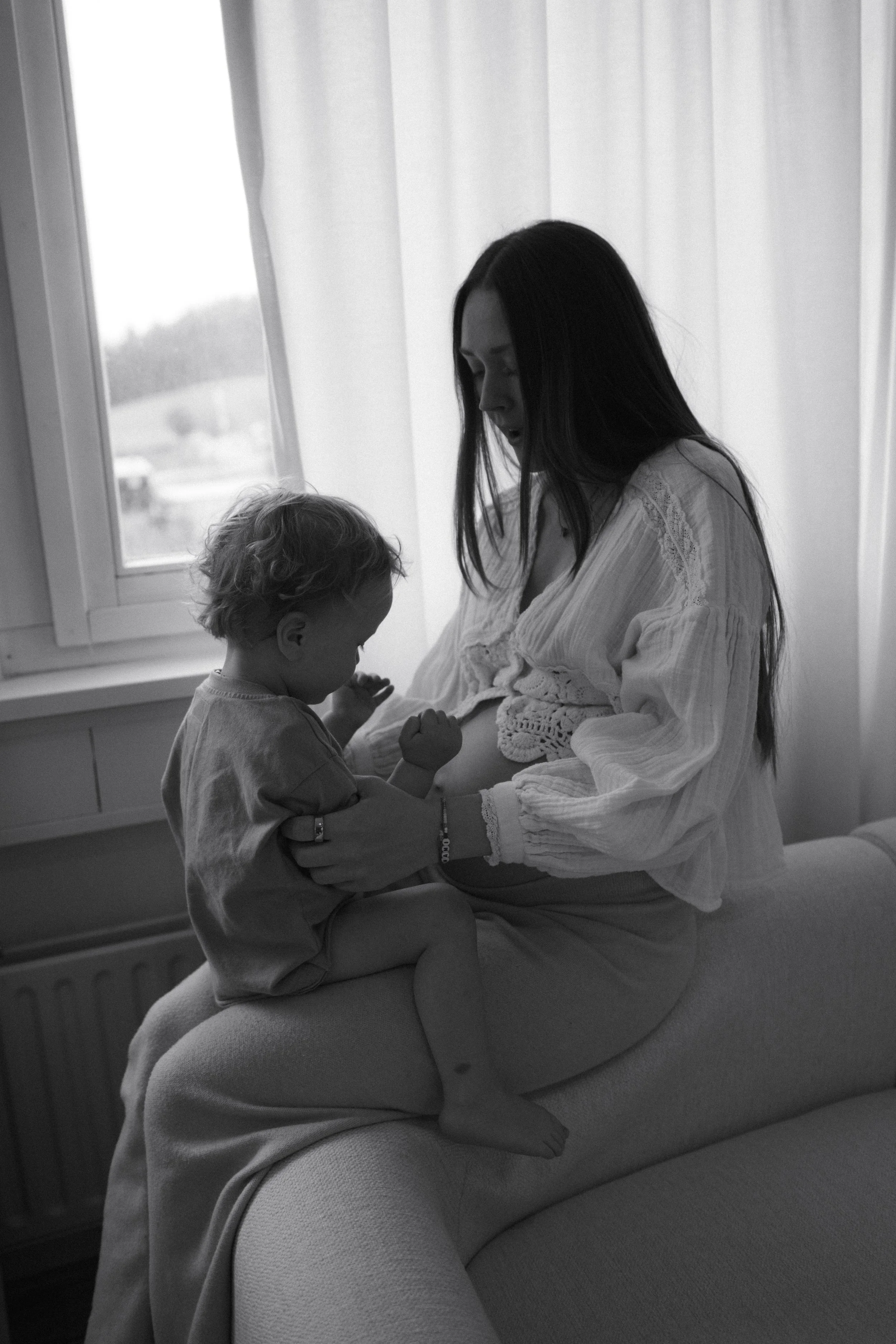 A pregnant woman sitting on a couch holding a young child in her lap while the child touches her pregnant belly, with a window and curtains in the background.