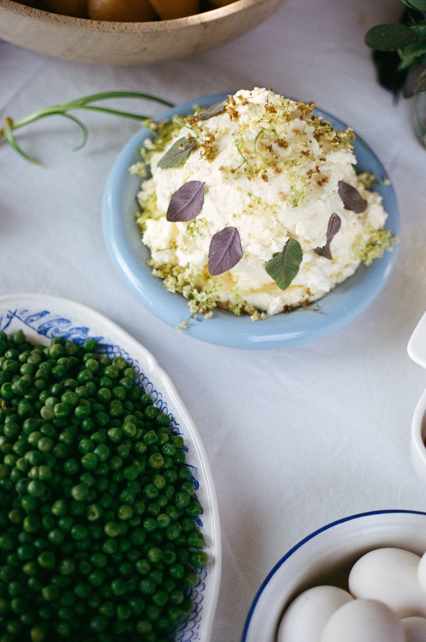 A bowl of potato salad garnished with herbs, a plate of green peas, and a bowl of white eggs.