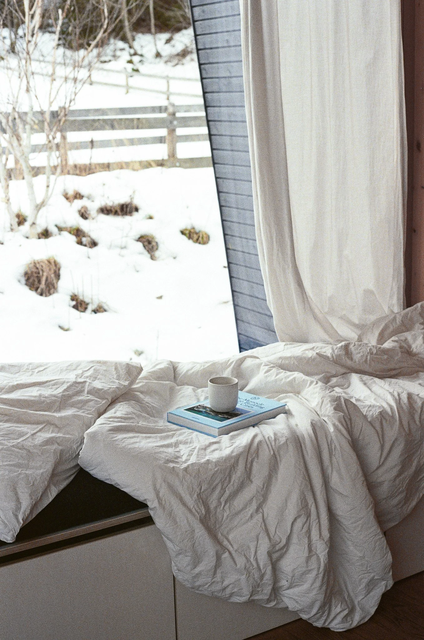 A bed with white rumpled bedding and a cup on top of a book, positioned in front of a large window showing a snowy outdoor landscape with trees and a fence.