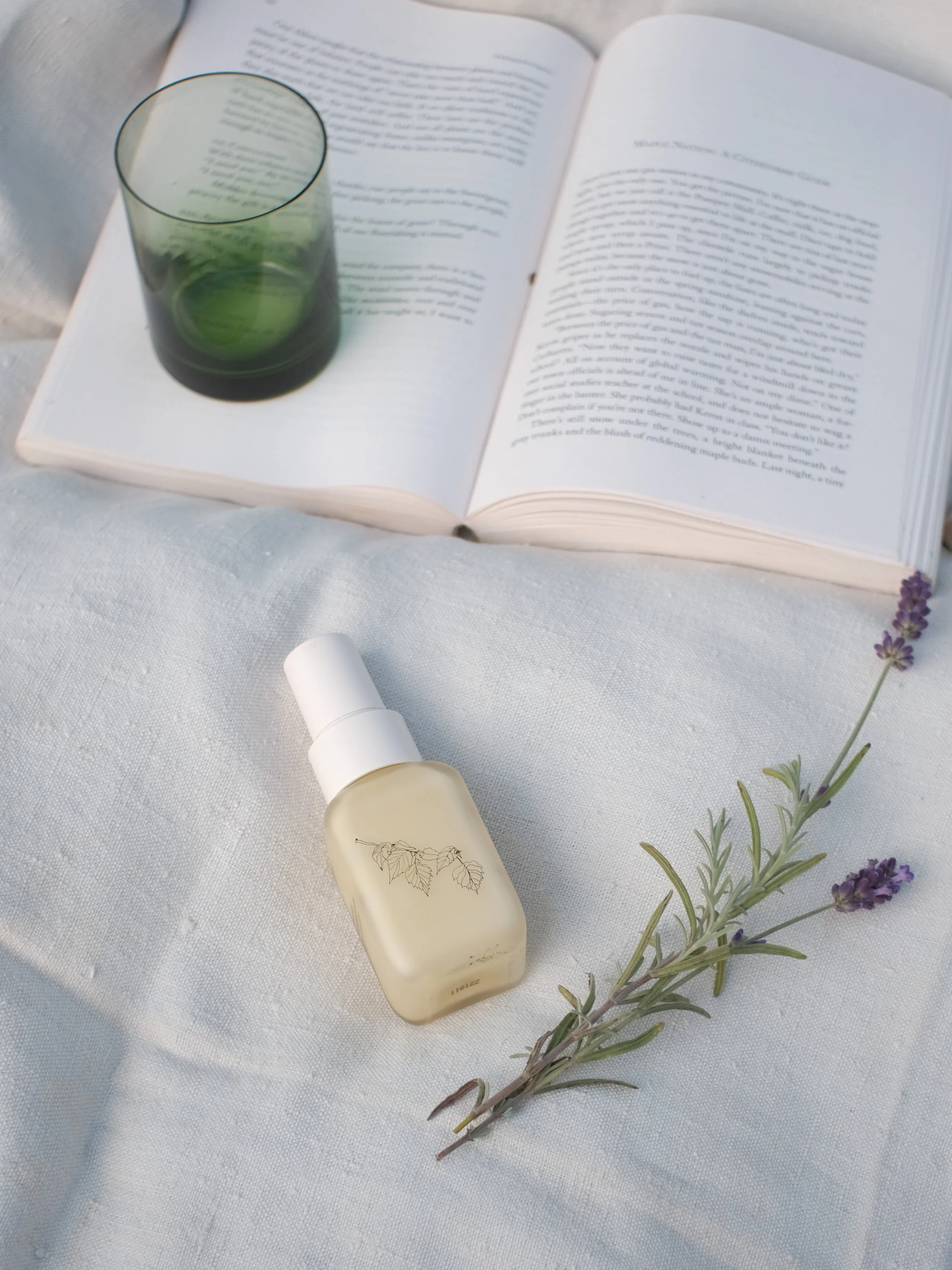 Open book with a green-tinted glass on top, a frosted spray bottle with a botanical illustration, and a sprig of lavender on a white fabric surface.