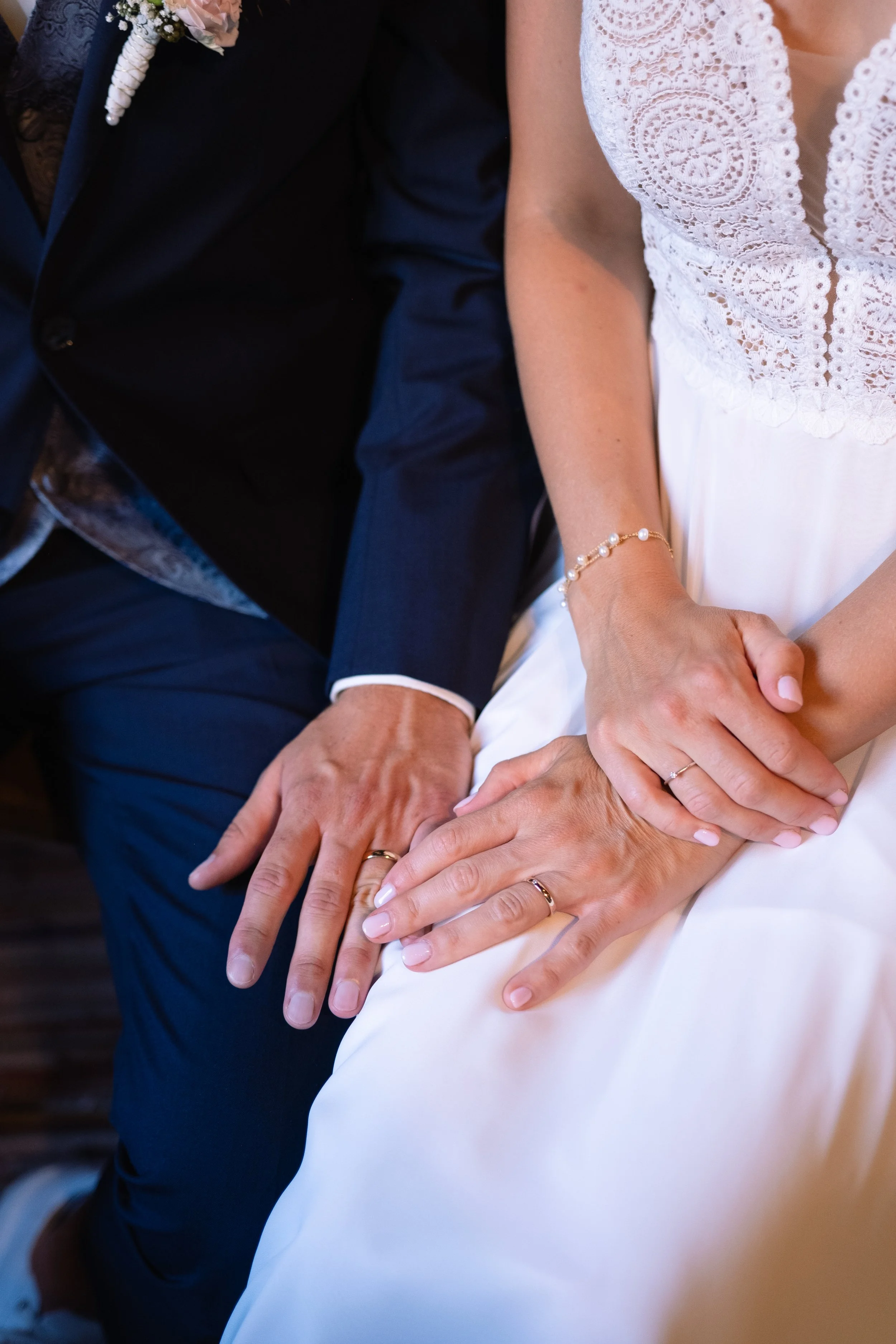 Close-up of a man and woman holding hands, both wearing wedding rings, with the woman in a white dress with lace details and the man in a dark suit.