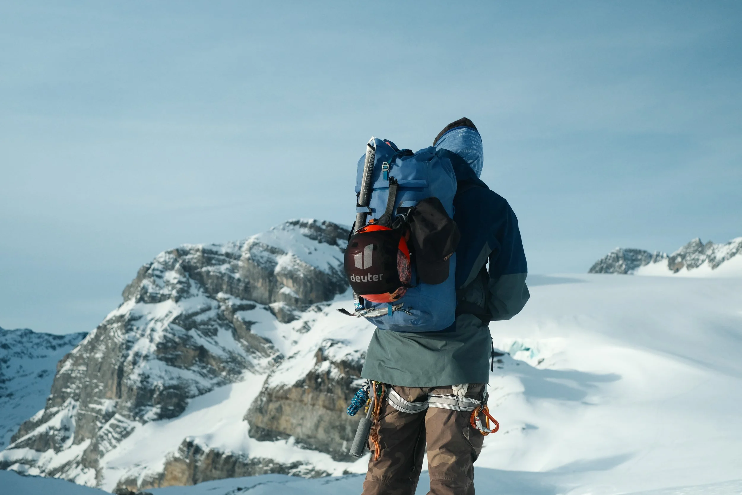 A person dressed in winter outdoor gear, standing on snow-covered mountain terrain with snow-capped peaks in the background.