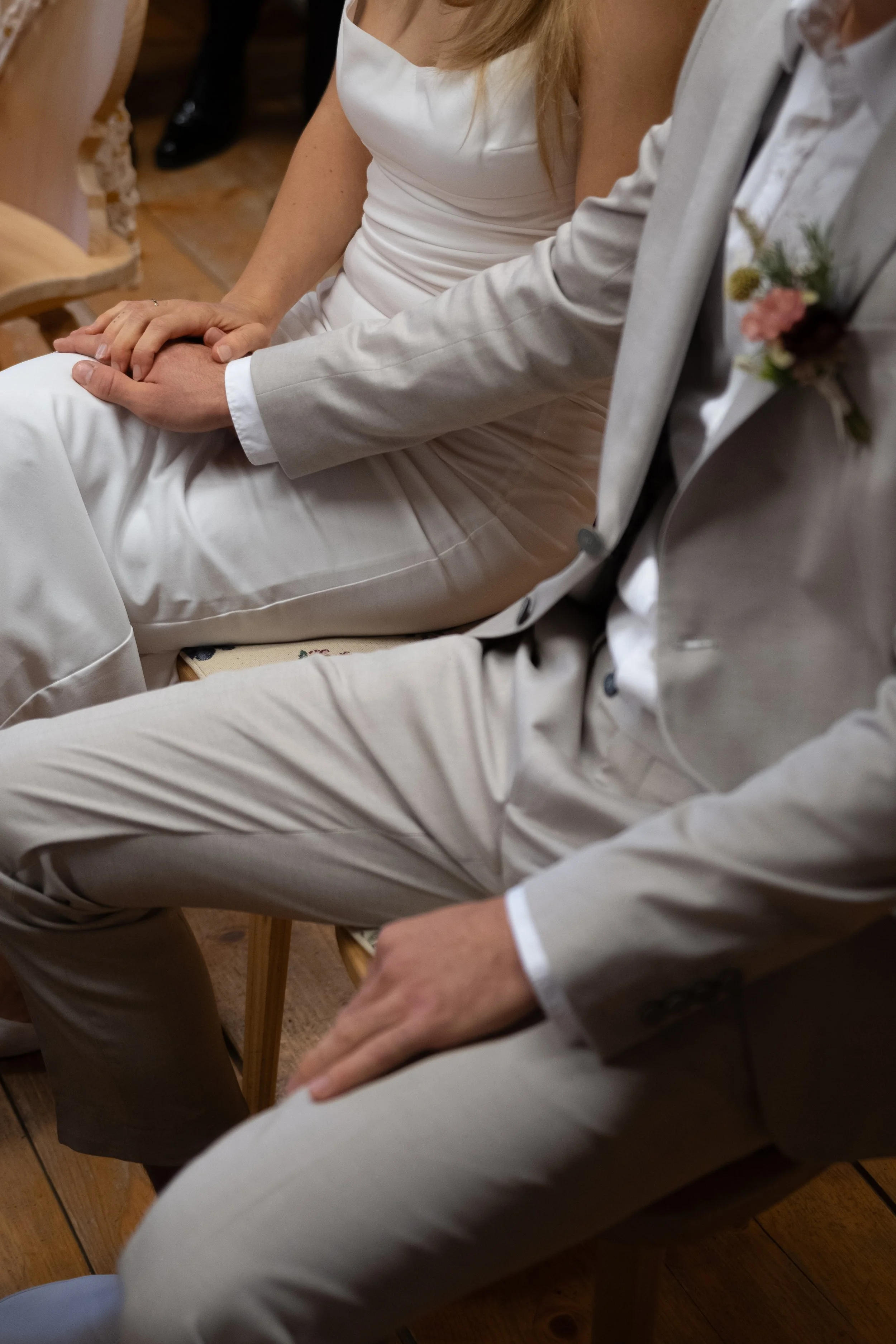 A bride and groom holding hands during a wedding ceremony, sitting on chairs inside a wooden room.