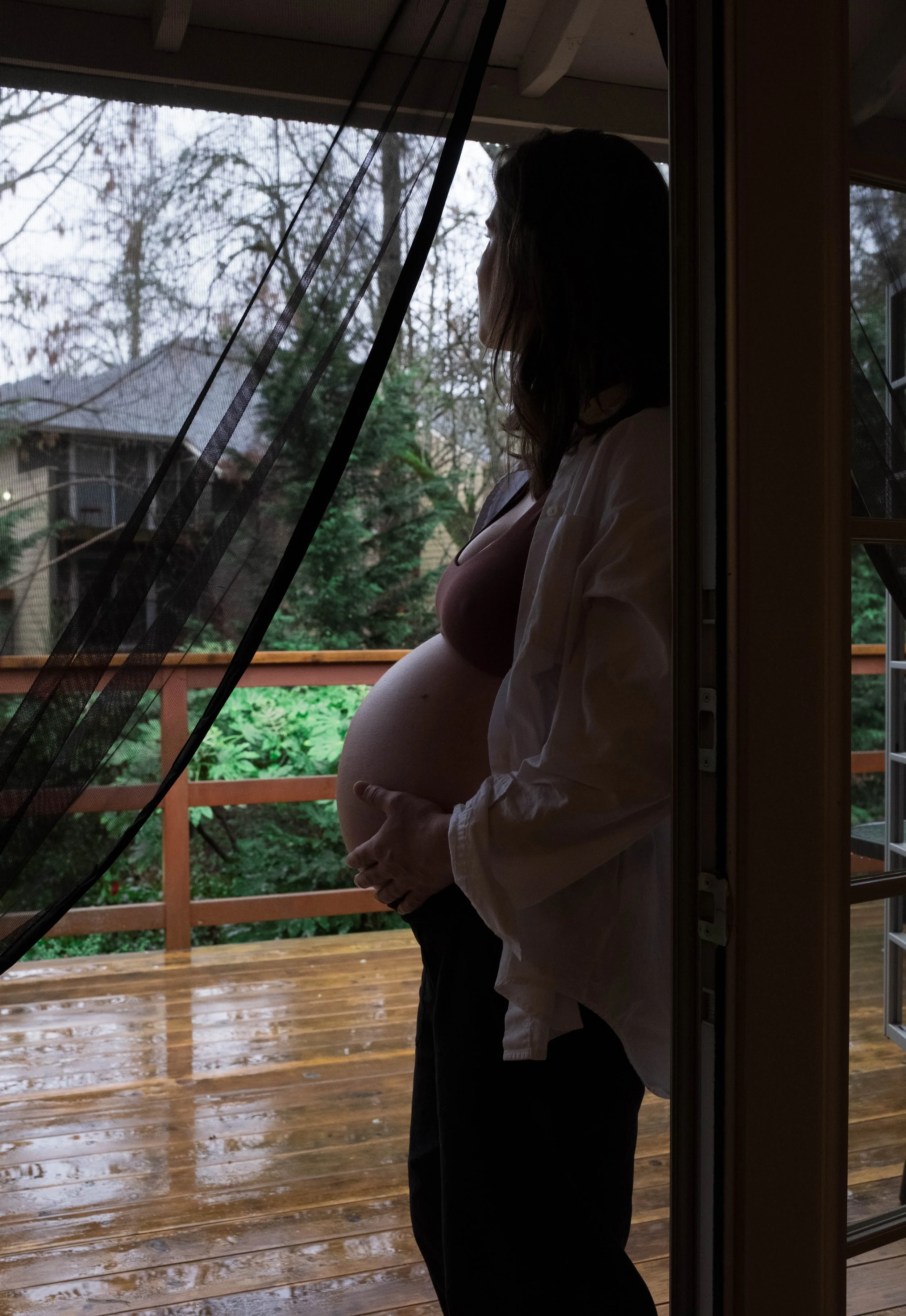 A pregnant woman standing by a doorway, looking outside on a rainy day, with her hand resting on her belly.