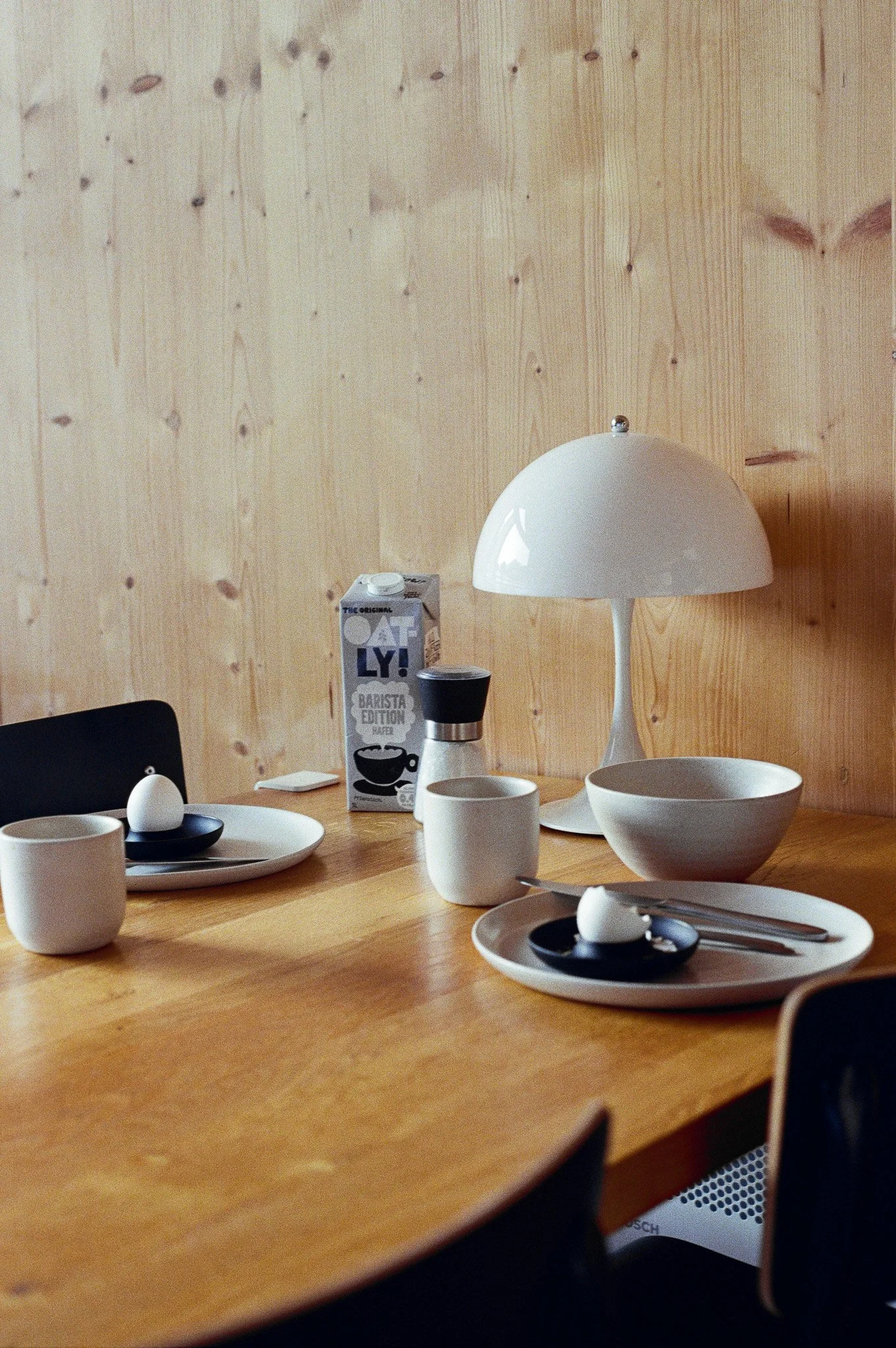 A wooden dining table set with white cups, black and white plates with boiled eggs, a carton of oat milk, a bowl, a plate, a lamp, a salt shaker, and some cutlery, against a wooden wall.