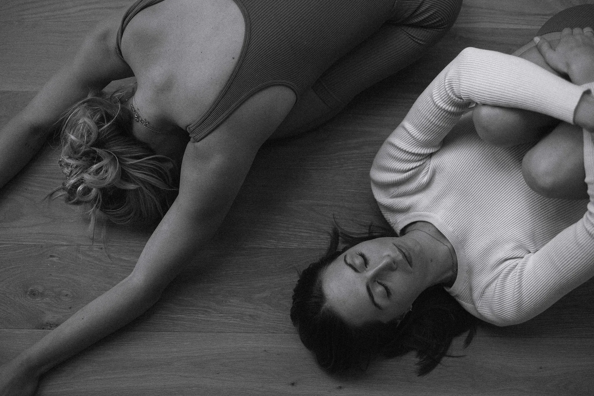 Two women lying on a wooden floor, one on her back with arms stretched out and the other curled up with knees drawn to her chest, both with eyes closed.