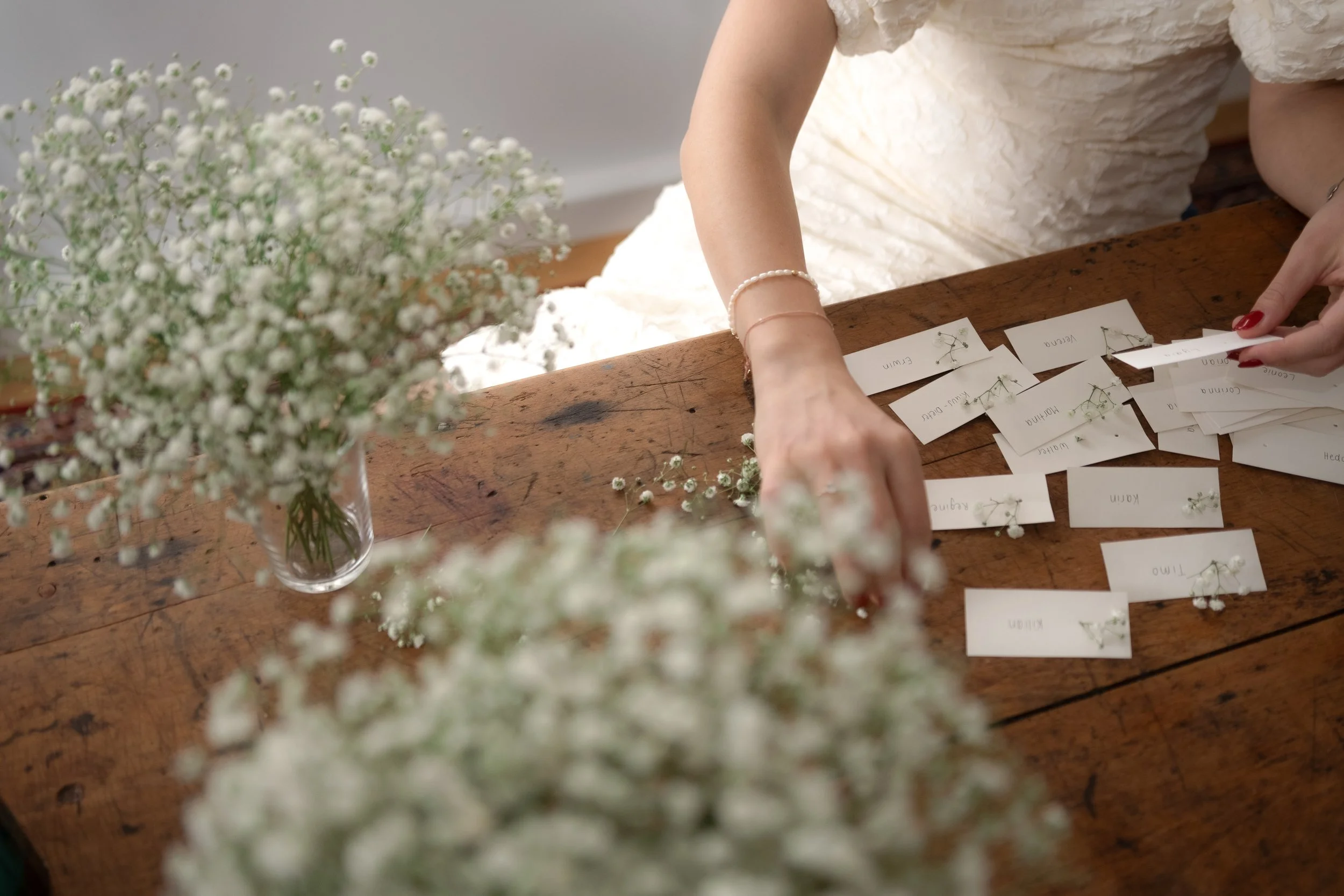 A woman assembling name tags with small white flowers on a wooden table, with a blurred bouquet of white flowers in a glass vase in the foreground.