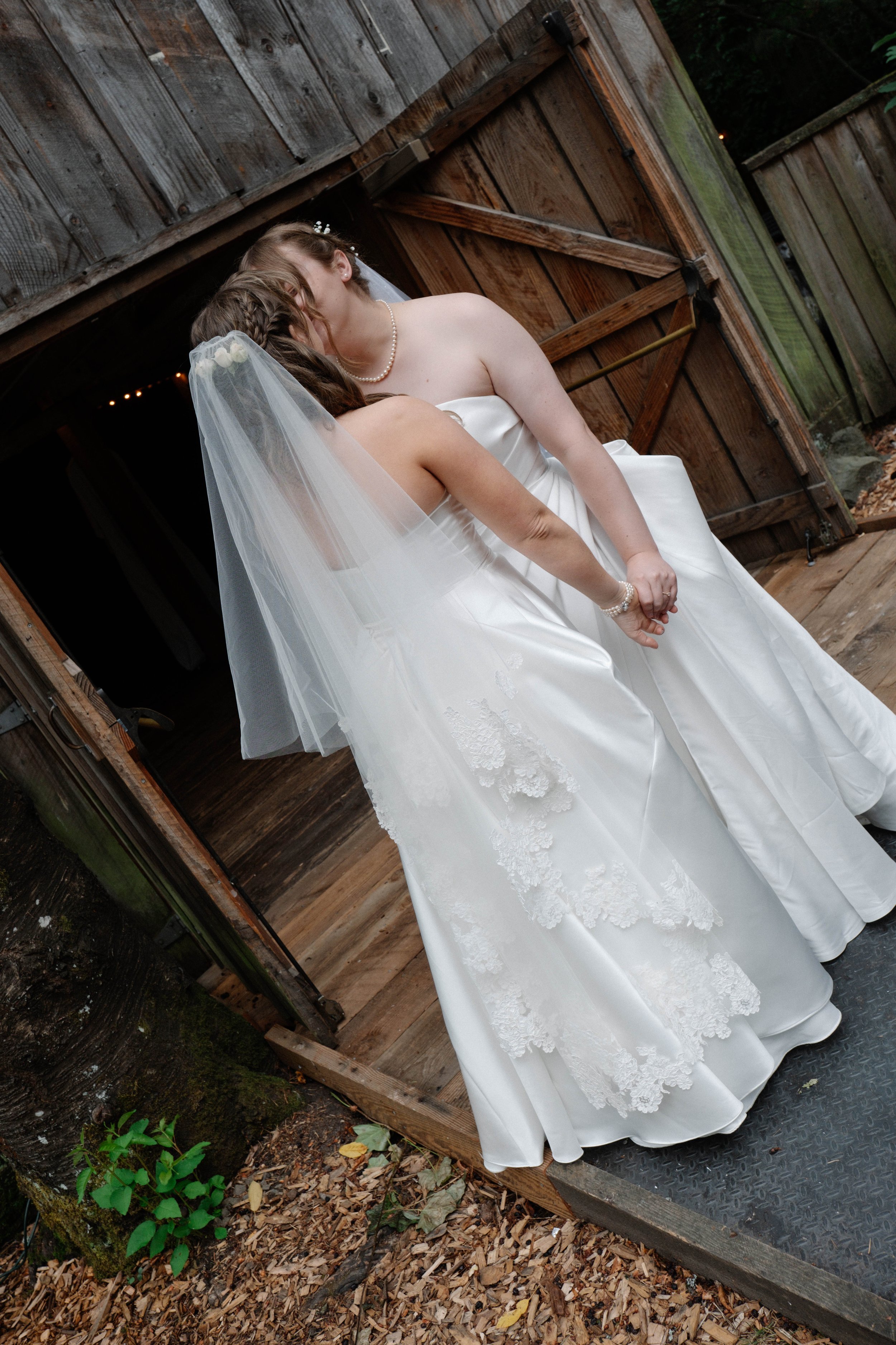 Two women dressed in wedding gowns sharing a kiss outdoors near a wooden shed in Washington and the Oregon coast.