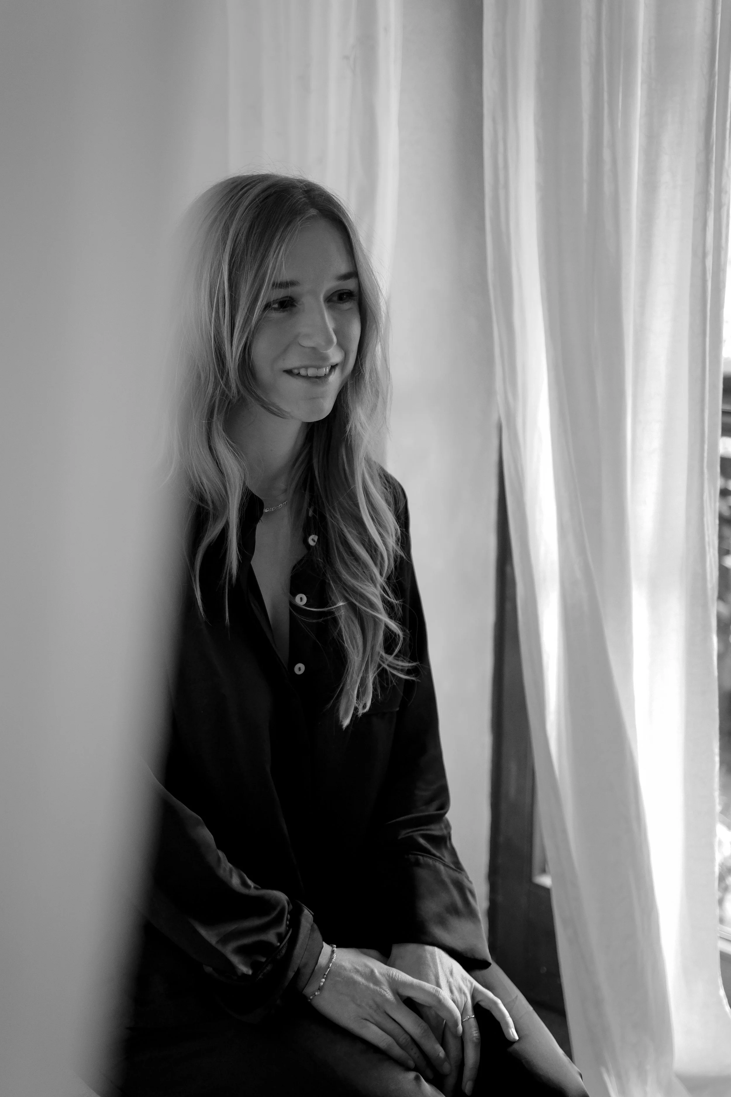 Black and white photo of a woman sitting by a window with curtains, smiling and looking to her left.