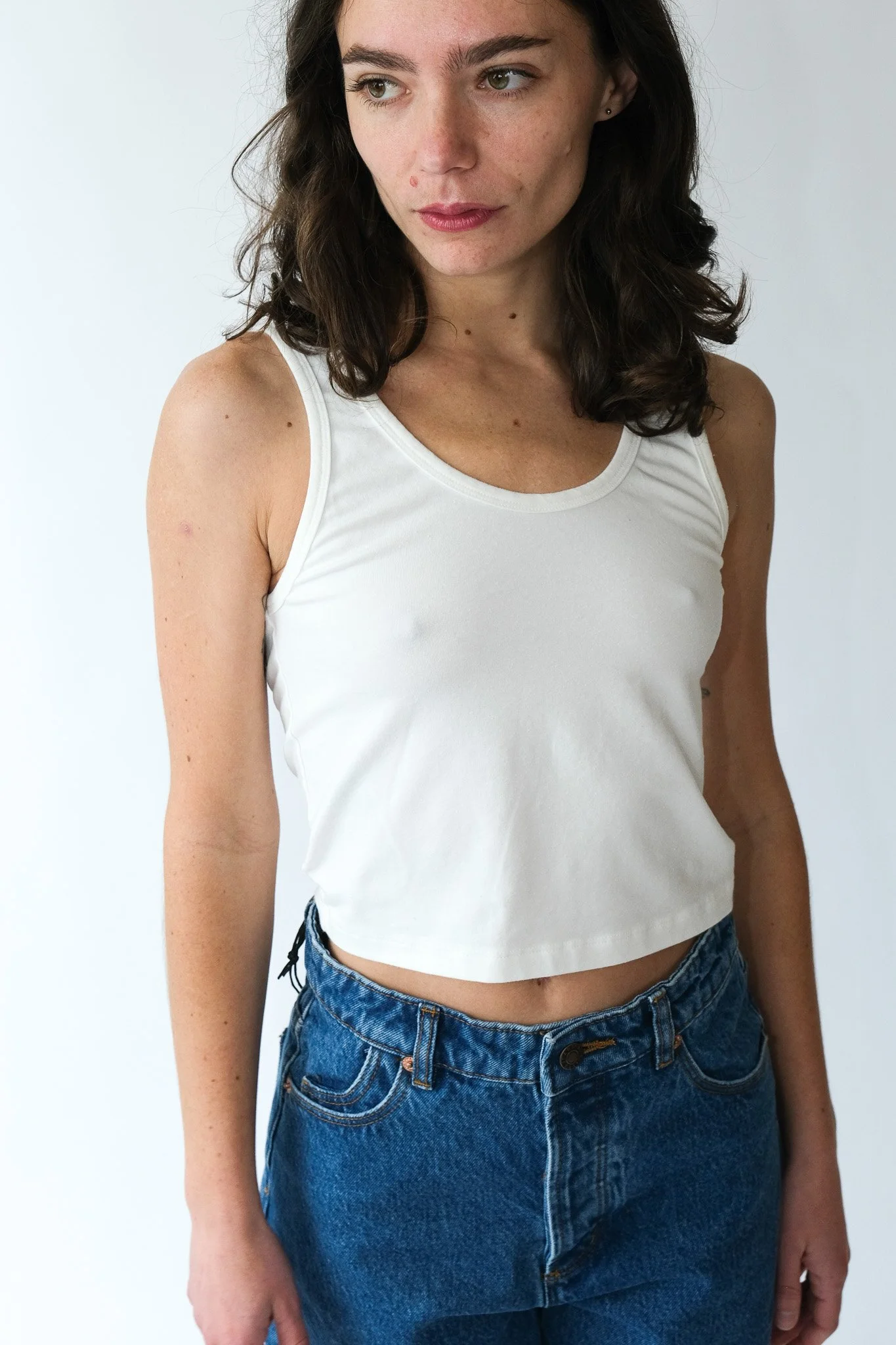 A young woman with dark curly hair wearing a white sleeveless crop top and blue jeans, standing against a plain light-colored background.