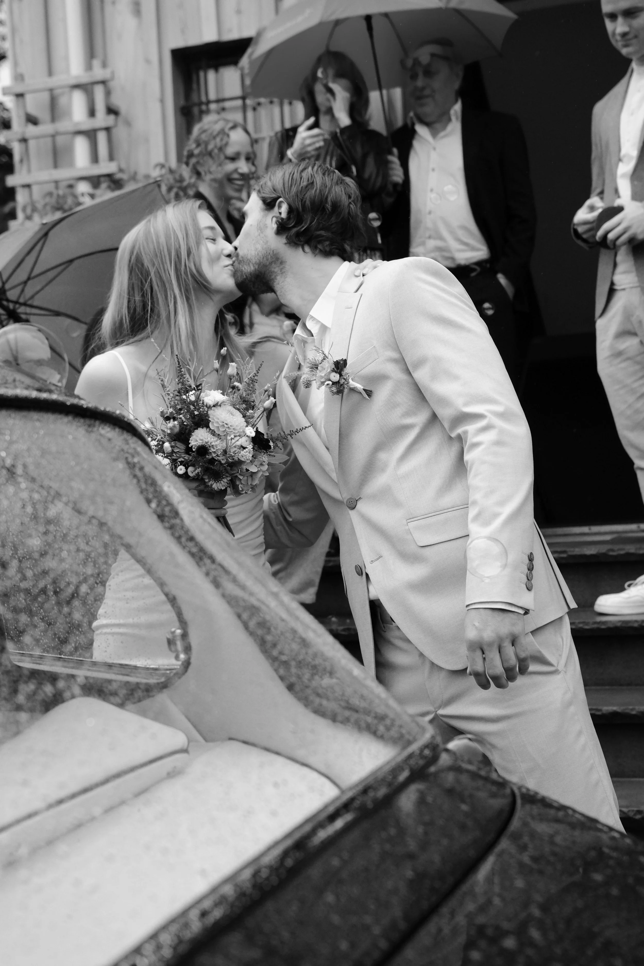 A couple sharing a kiss during their wedding, with the bride holding a bouquet of flowers, and guests with umbrellas in the background.