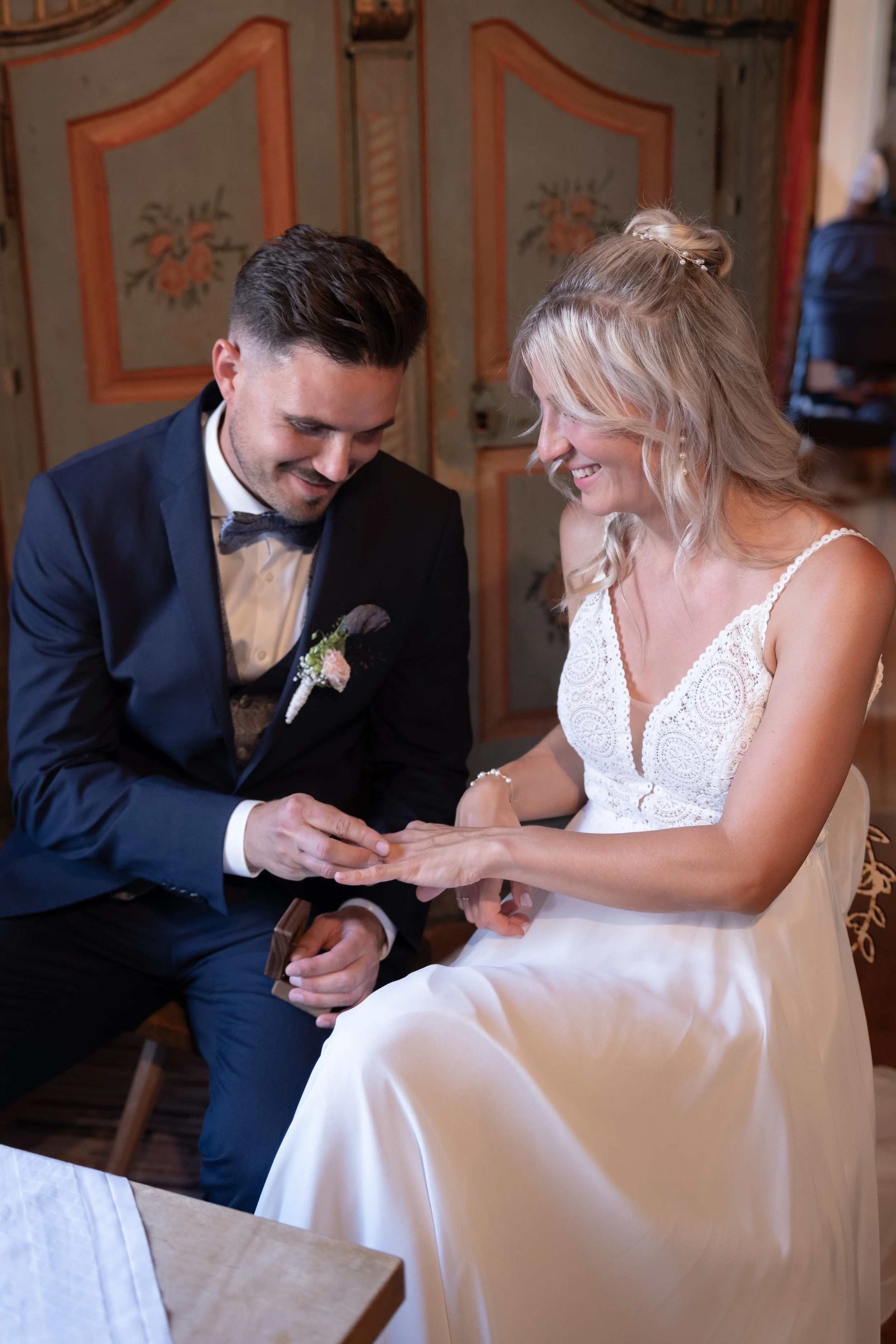 A groom placing a ring on a bride's finger during a wedding ceremony. The groom is wearing a navy suit with a bow tie, and the bride is dressed in a white lace wedding gown. They are smiling and appear happy.