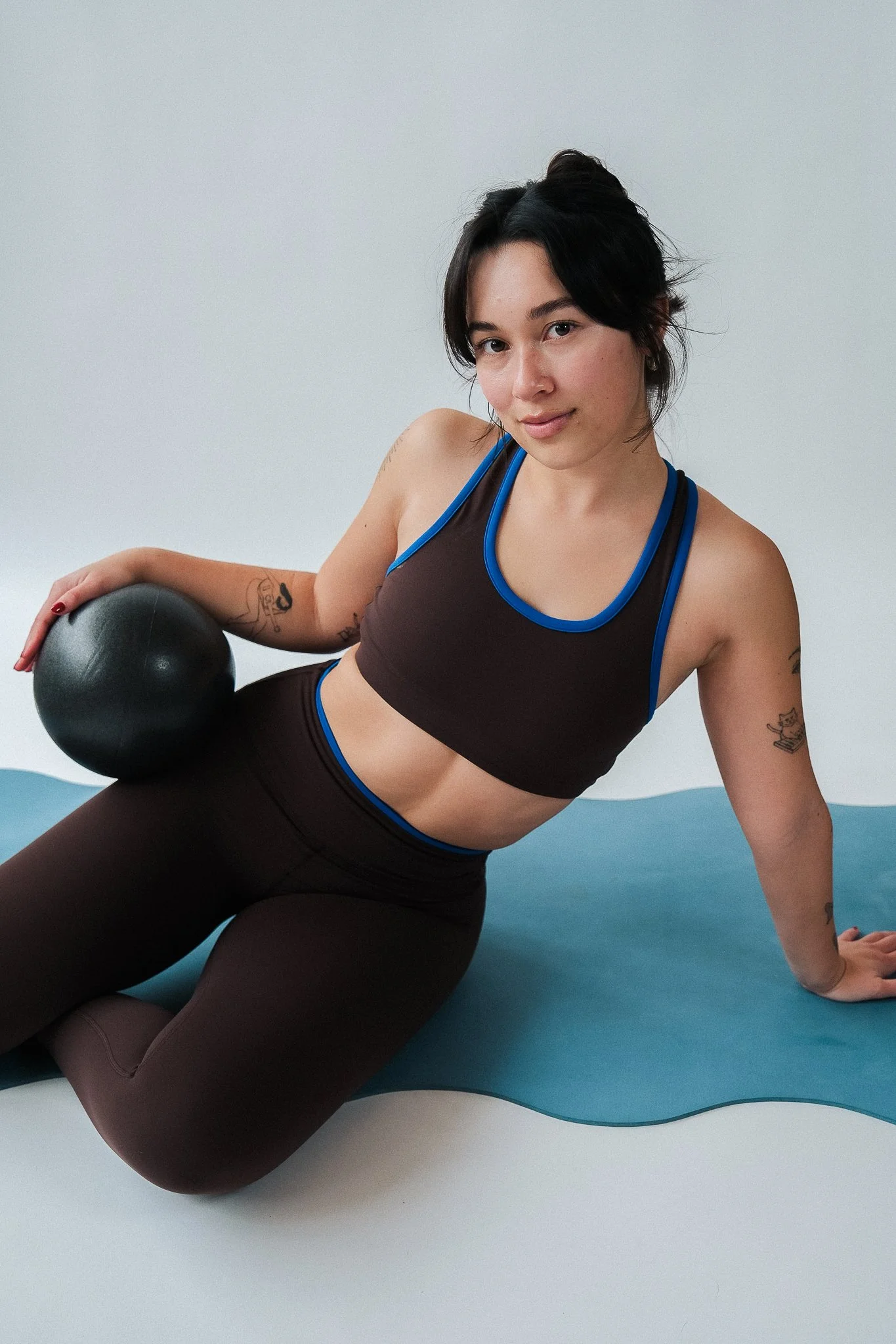 A woman in workout attire holding a black medicine ball, sitting on a yoga mat, against a plain white background.