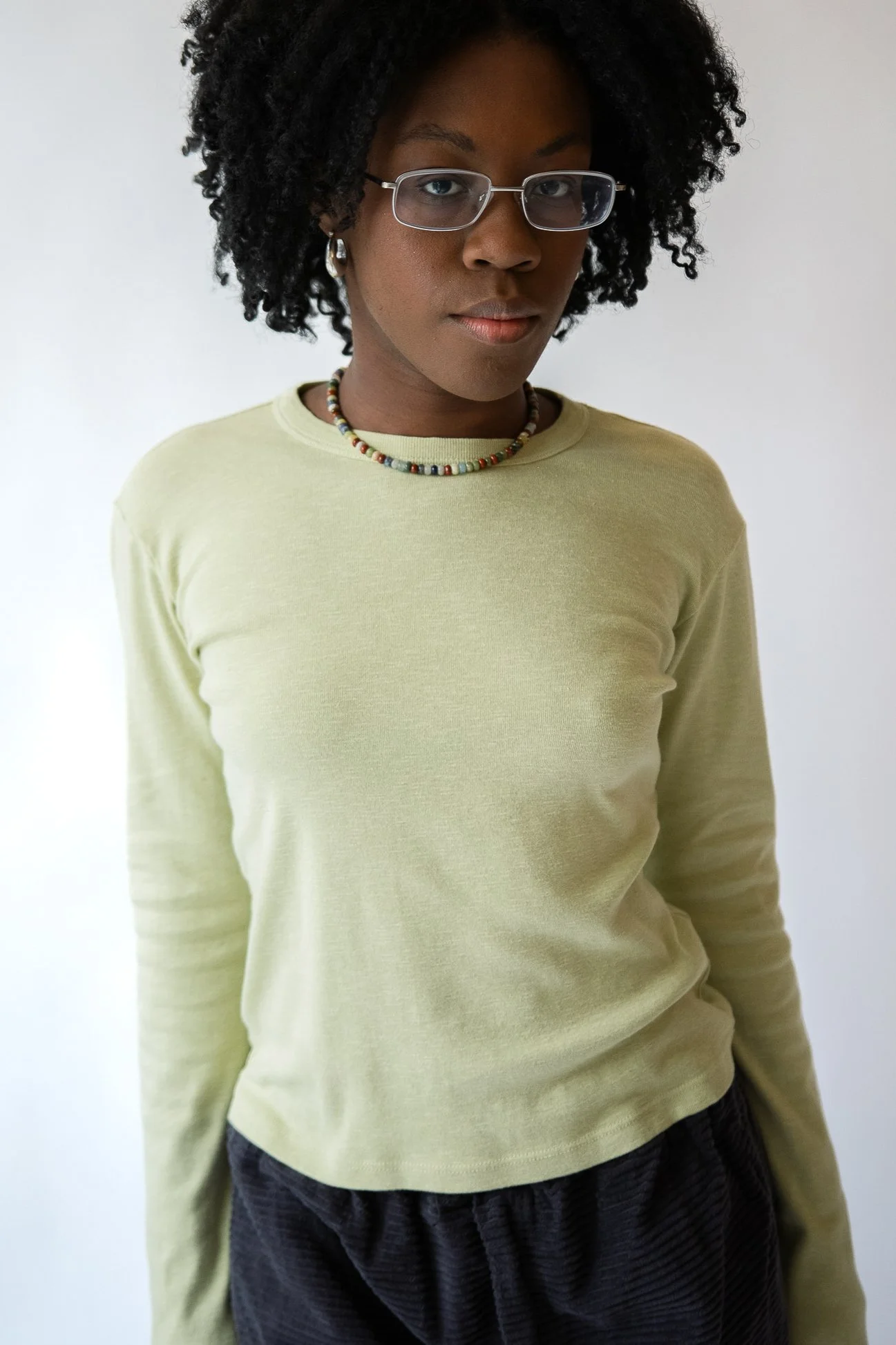 Portrait of a young woman with dark curly hair, glasses, wearing a light yellow long-sleeve shirt, black textured pants, and a colorful beaded necklace, standing against a plain white background.