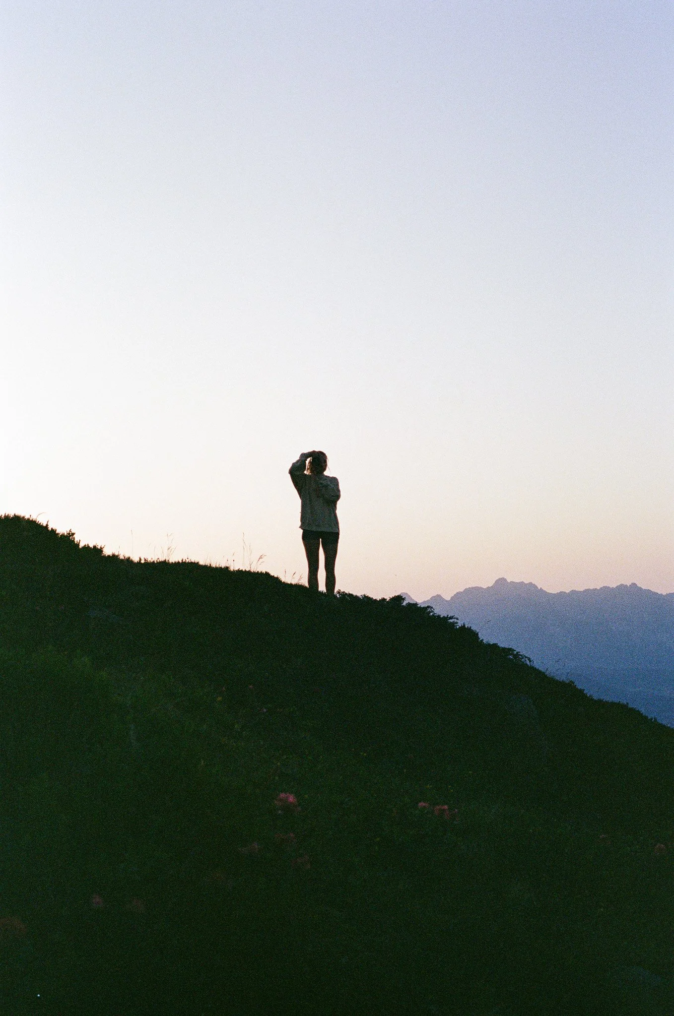 Silhouette of a person standing on a hill at dusk, looking towards the horizon with distant mountains in the background.