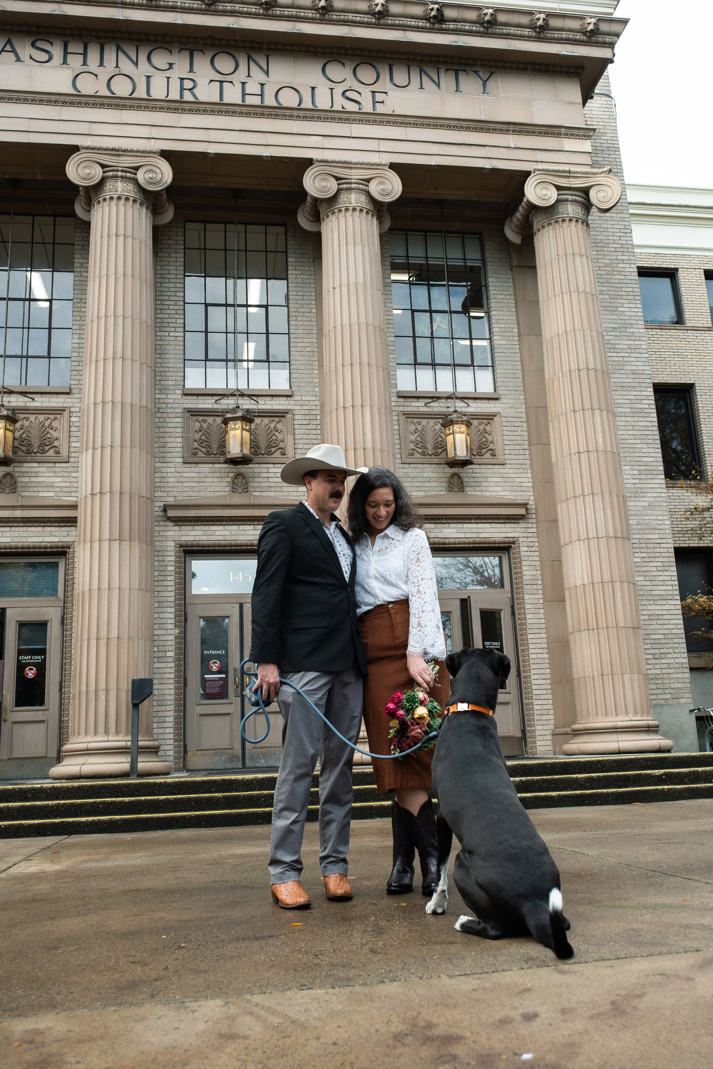 A couple stands in front of the Washington County Courthouse, smiling with their dog holding a bouquet of flowers. The courthouse features large columns and decorative lanterns.