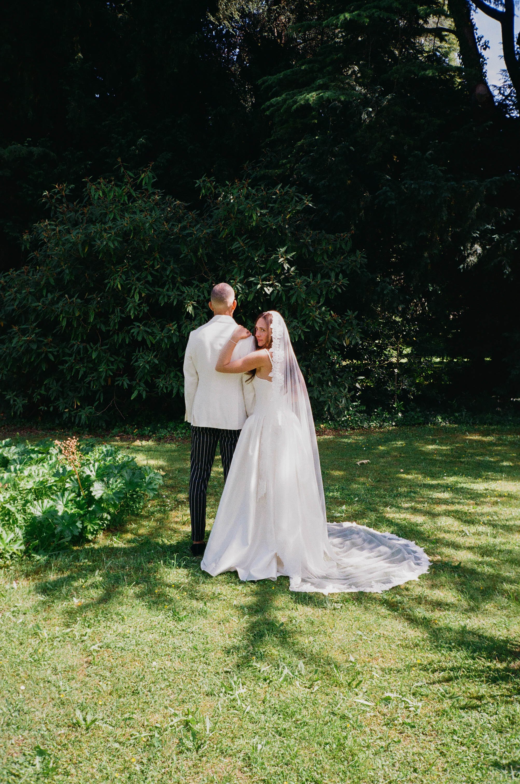 A bride and groom standing in a grassy garden area with lush green trees in the background. The bride is wearing a white wedding gown and veil, and the groom is dressed in a white blazer with black and white striped pants. They are embracing, with th