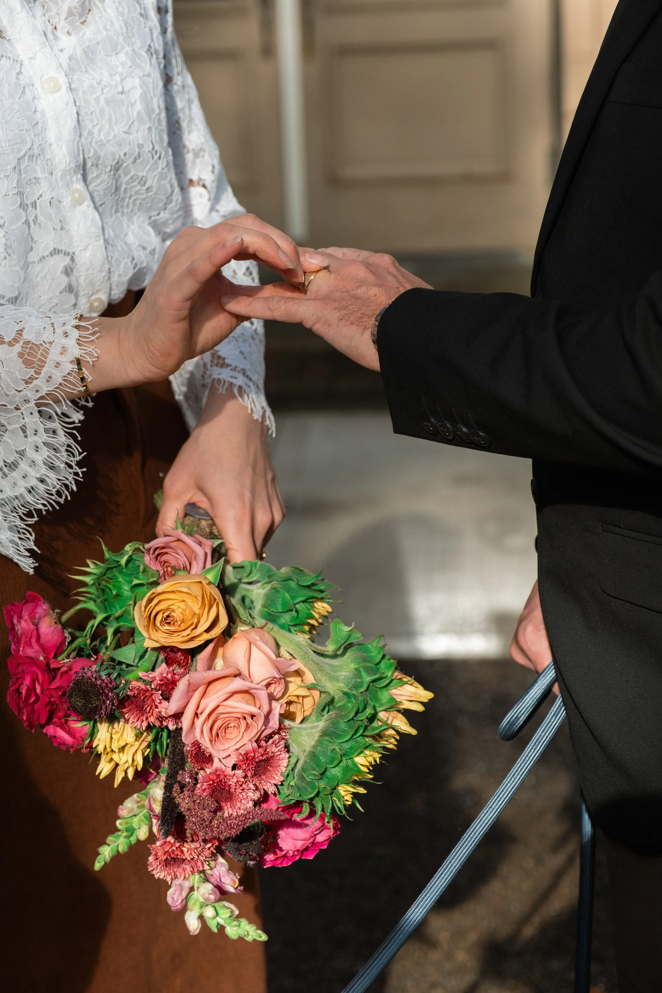 A couple exchanging rings during a wedding ceremony. The woman is holding a vibrant bouquet of pink, orange, and yellow roses with greenery, while the man is dressed in a black suit. They are indoors, and the focus is on the hands and bouquet.