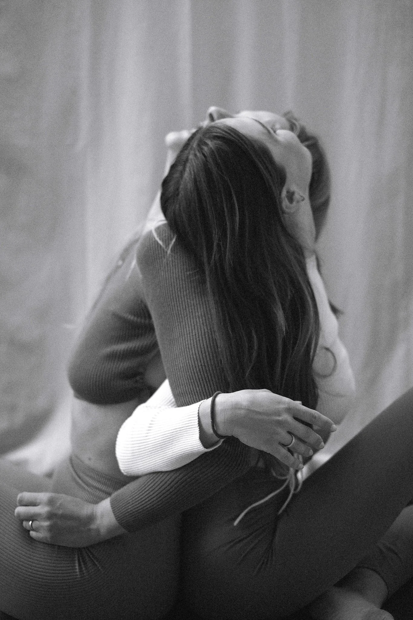 Black and white photo of a woman with long hair, leaning back with her head tilted upwards, eyes closed, and a hand resting on her chest, sitting against a curtain backdrop.